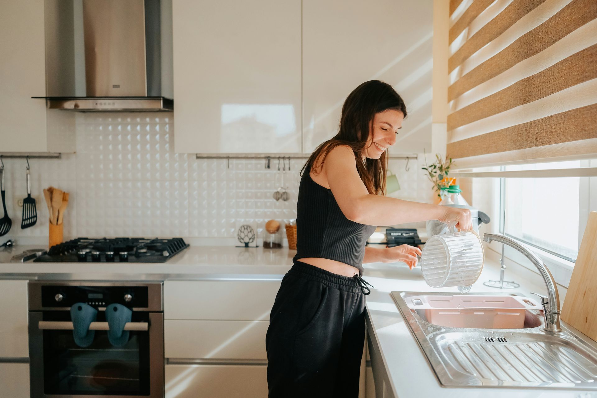 Woman filling a water bottle at a kitchen sink, smiling. Sunny kitchen with white cabinets.