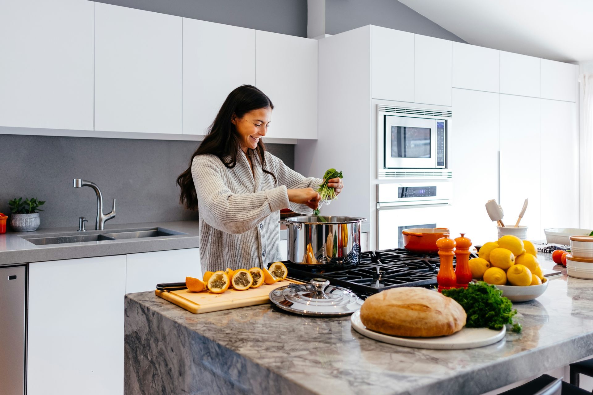 Woman adds greens to a pot on a stovetop in a bright kitchen with a granite island, bread, and lemons.