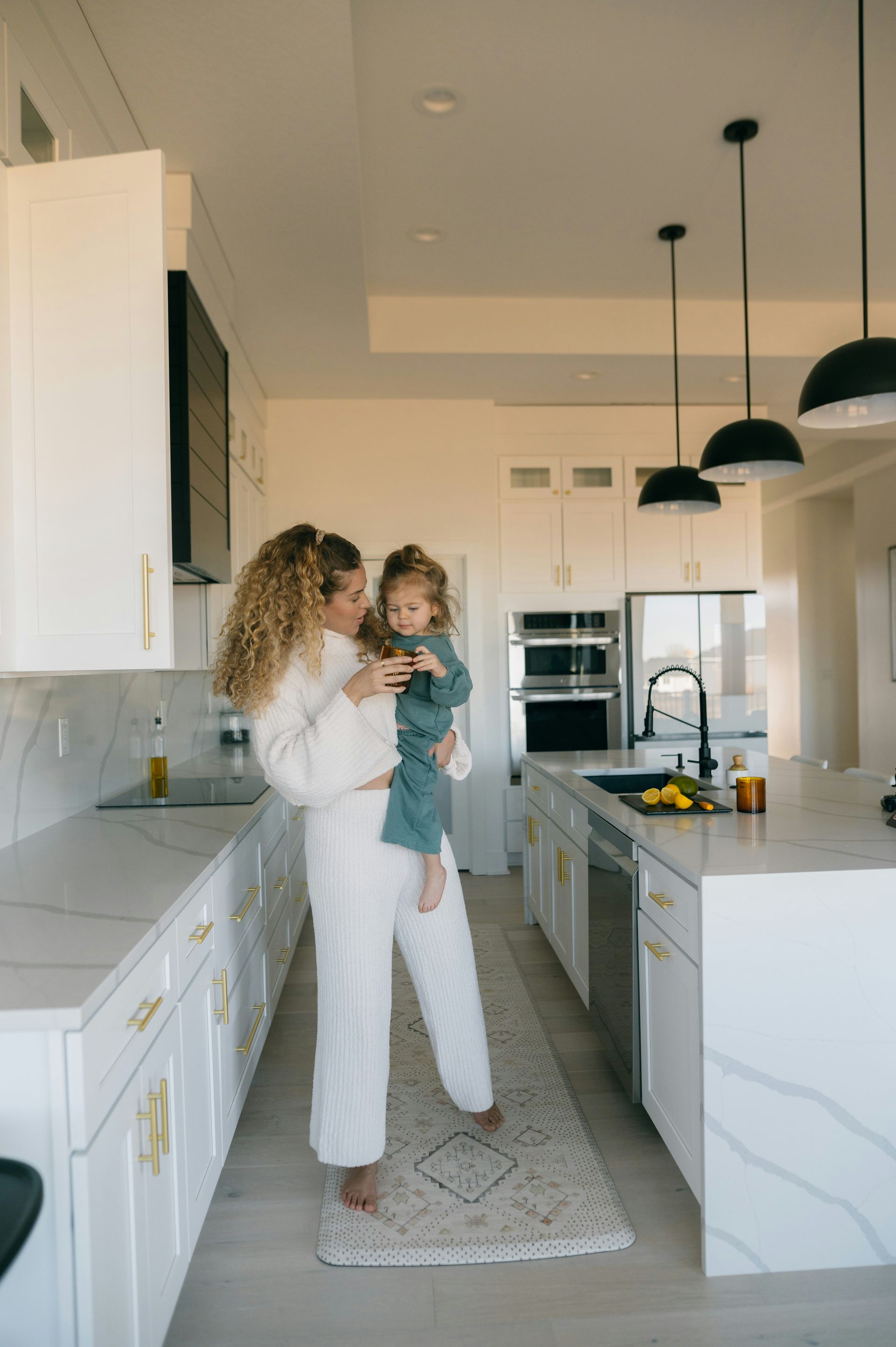 Woman holding a child in a bright white kitchen. The woman smiles, looking at the child.