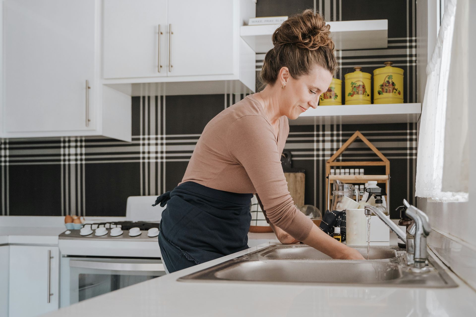 Woman washing dishes in a kitchen. She wears an apron, has her hair up, and is smiling.