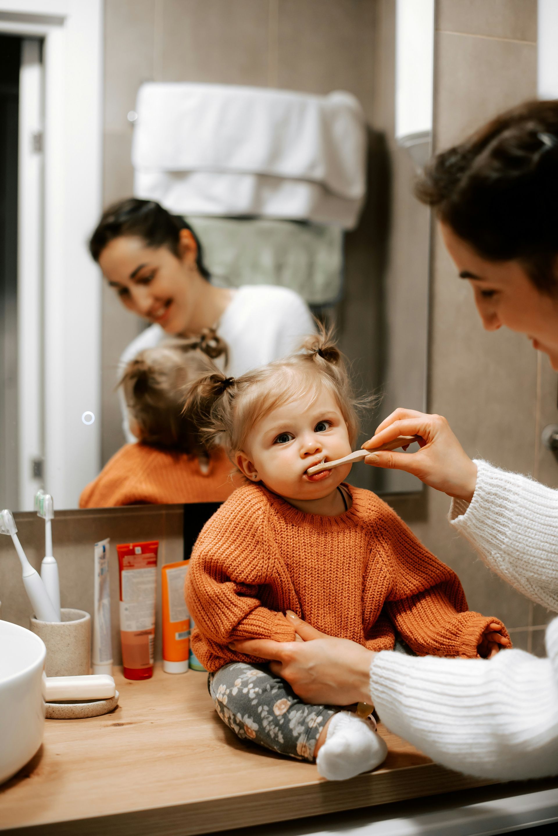 Woman brushing a toddler's teeth in a bathroom; child sits on counter, orange sweater.