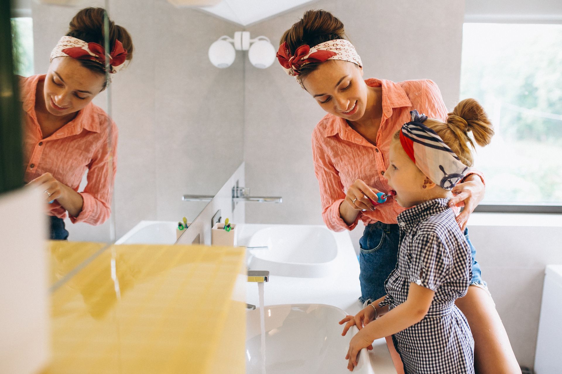Woman helping child brush teeth in bathroom. Smiling, near sink and mirror.