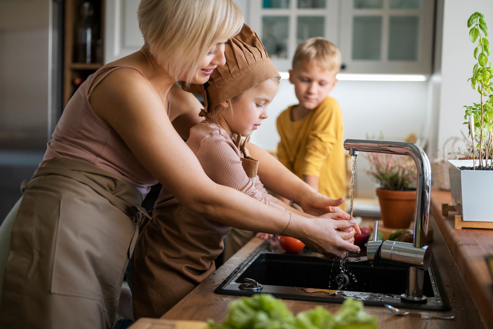 Woman helps child wash produce in kitchen sink; another child watches.
