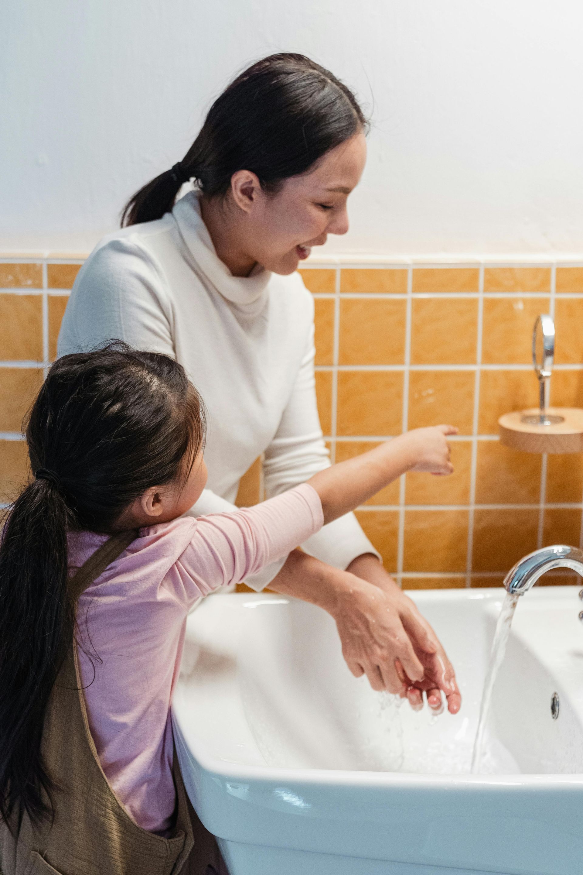 Woman and child washing hands together in a bathroom, water running from the faucet.
