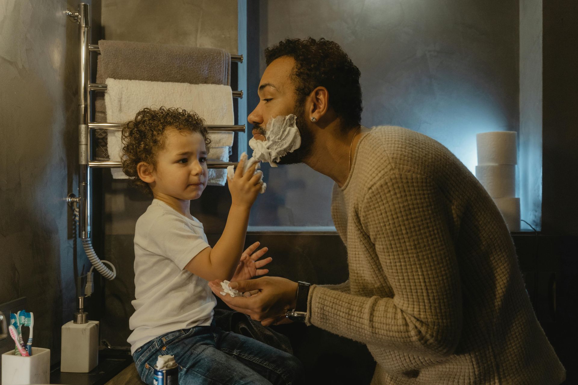 Father and child in bathroom; child playfully applies shaving cream to father's face.