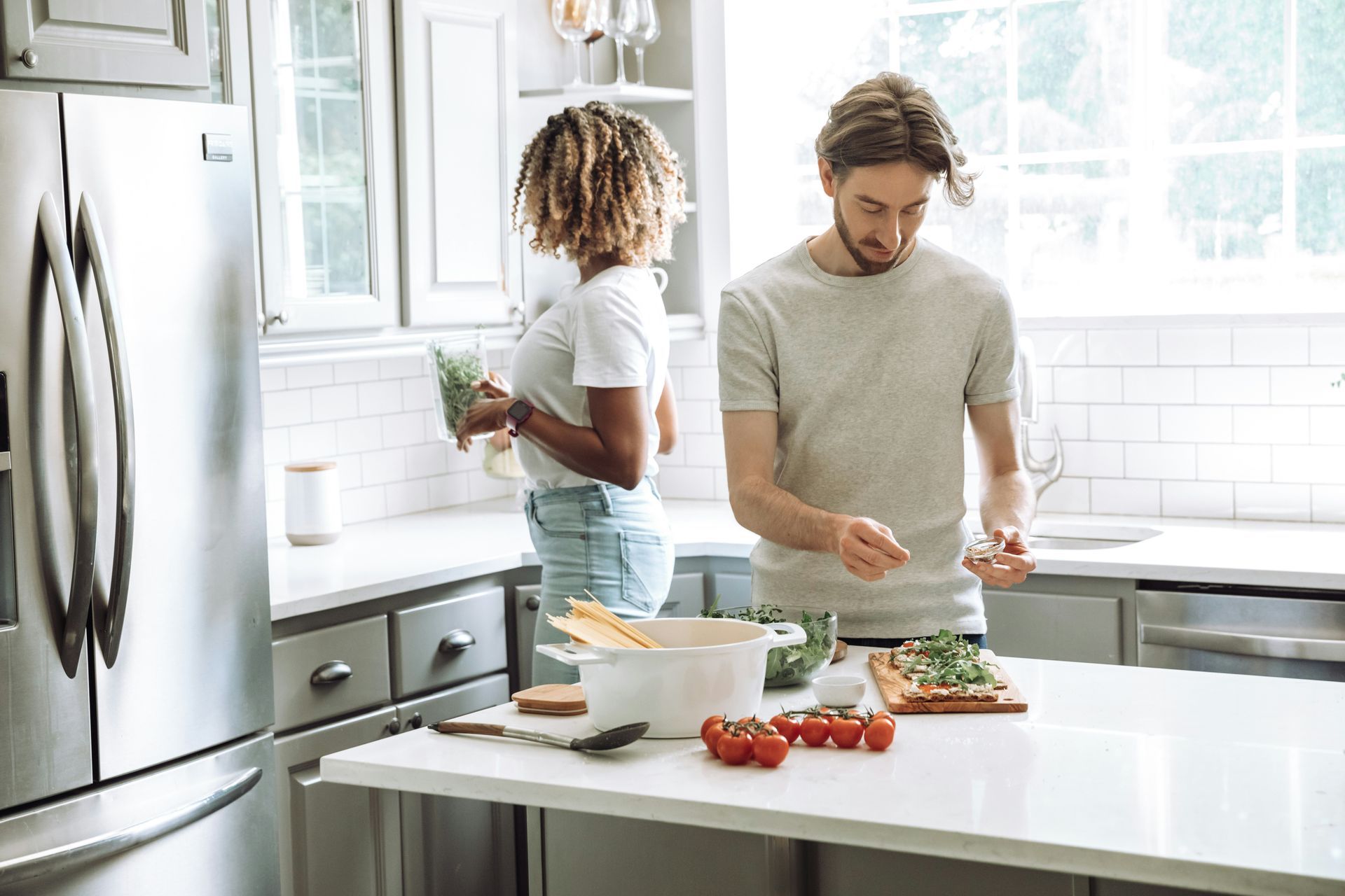 Couple cooking in a bright kitchen: man chopping herbs, woman holding greens, pasta and tomatoes on the counter.