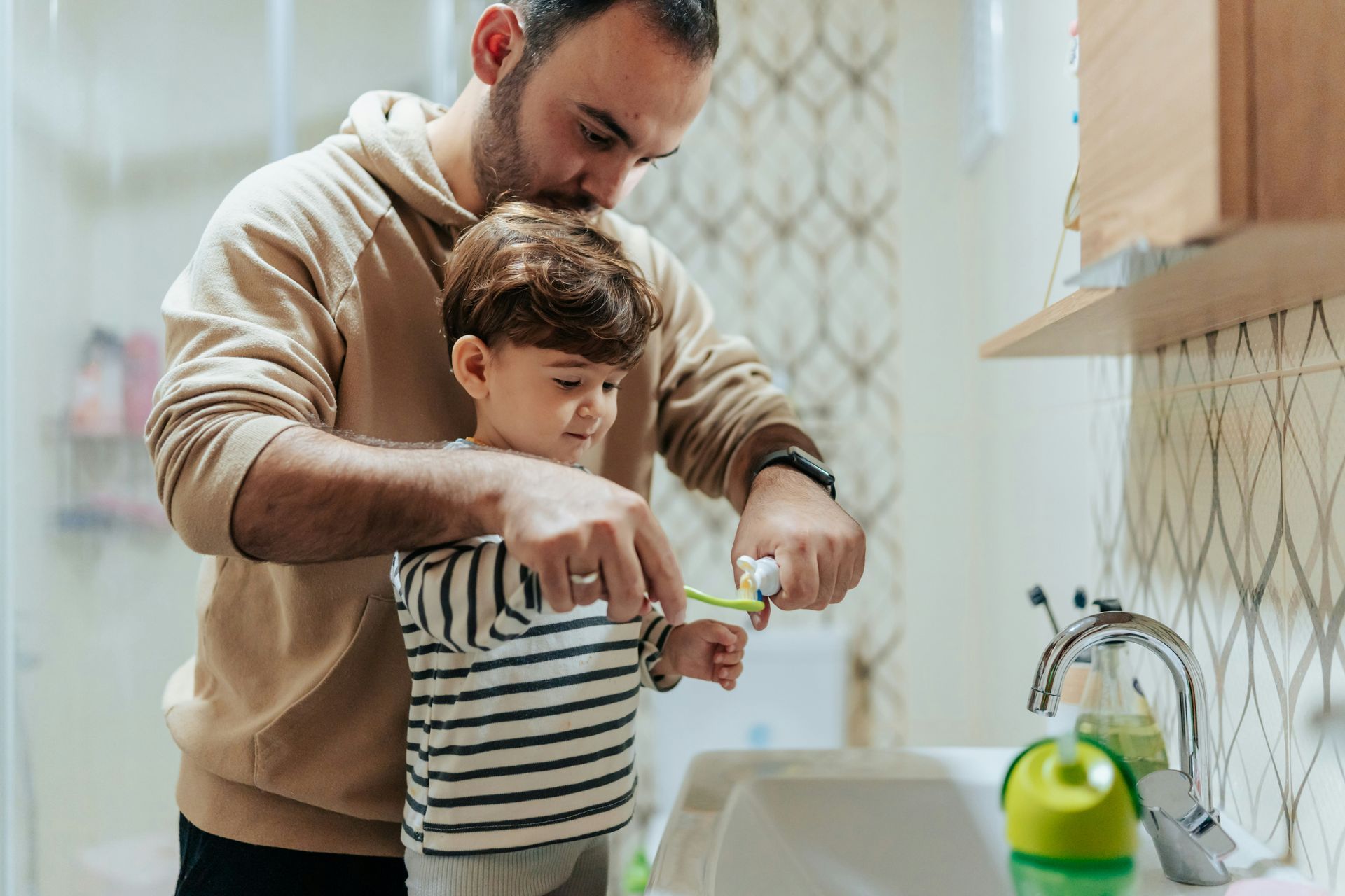 Man helping a child brush their teeth in a bathroom; applying toothpaste to a toothbrush.