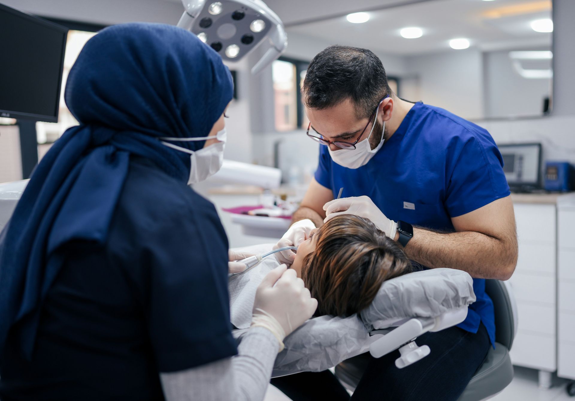 Dentist and assistant examining a patient's teeth in a dental clinic; patient is in a dental chair.