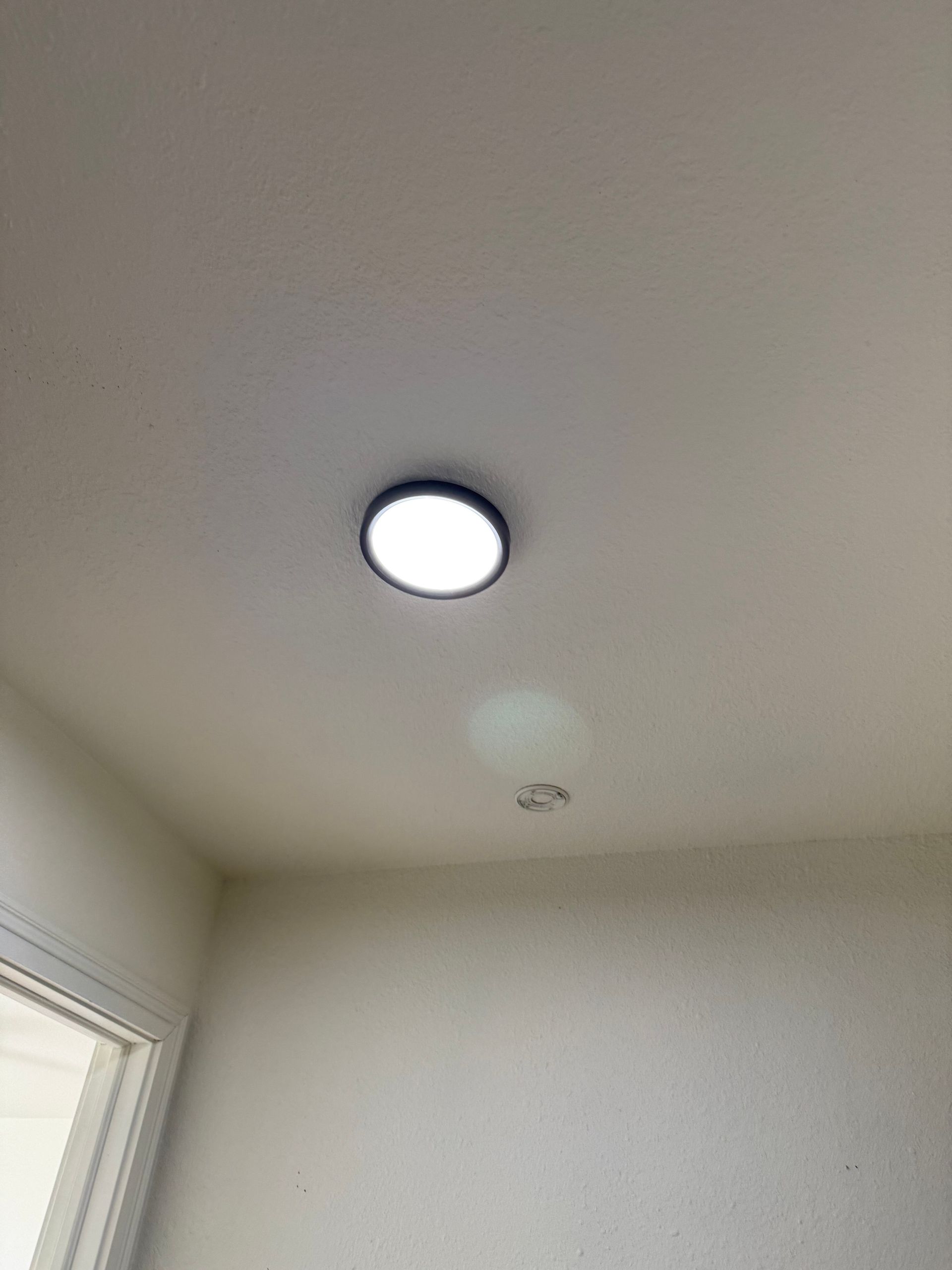 Ceiling corner with a round flush-mounted light and a window edge in a white room.