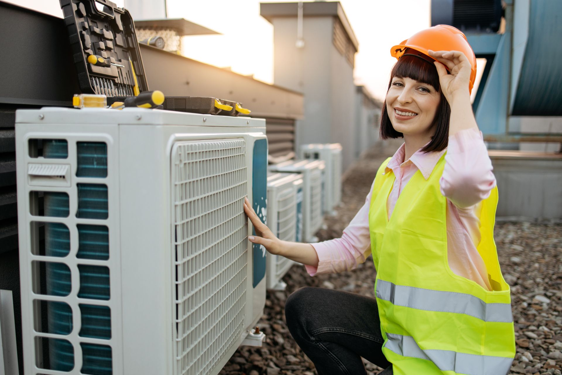 A woman in a yellow vest and hard hat is working on an air conditioner.