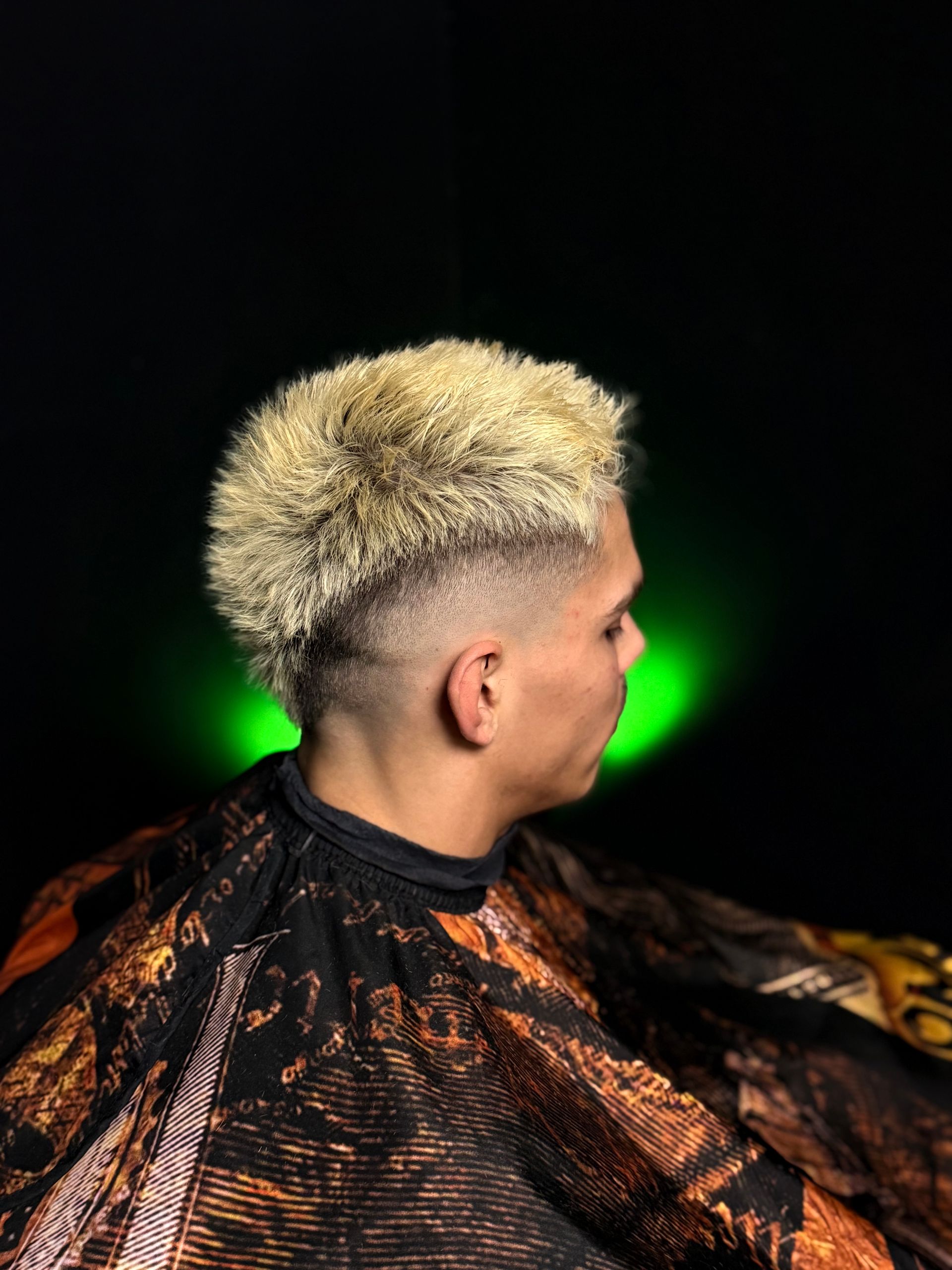 Person with a bleached, spiky hairstyle, skin faded from dark to light. Black and orange cape, green light in background.