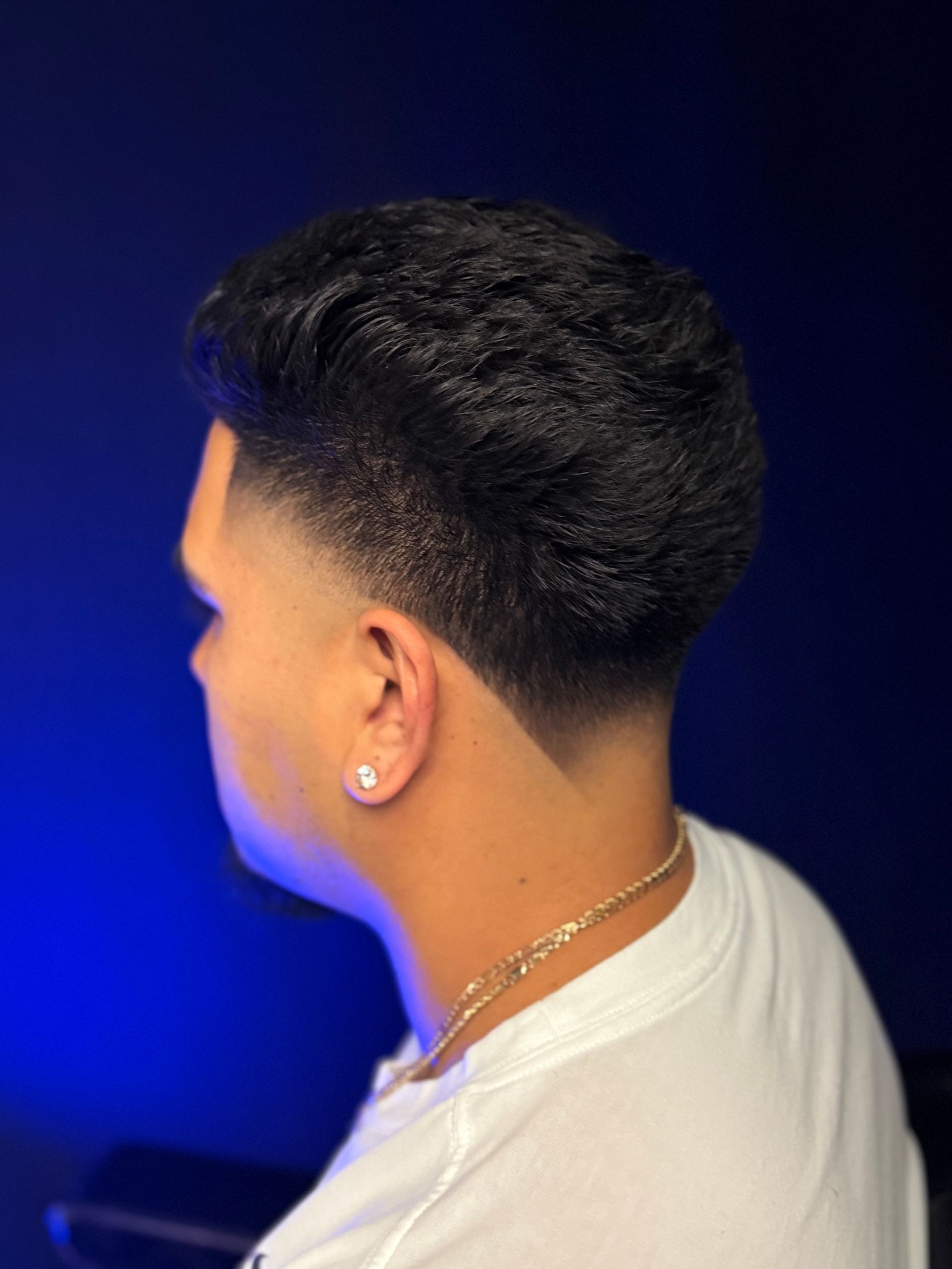 Man with a low fade haircut, dark hair, wearing a white shirt, against a blue background.