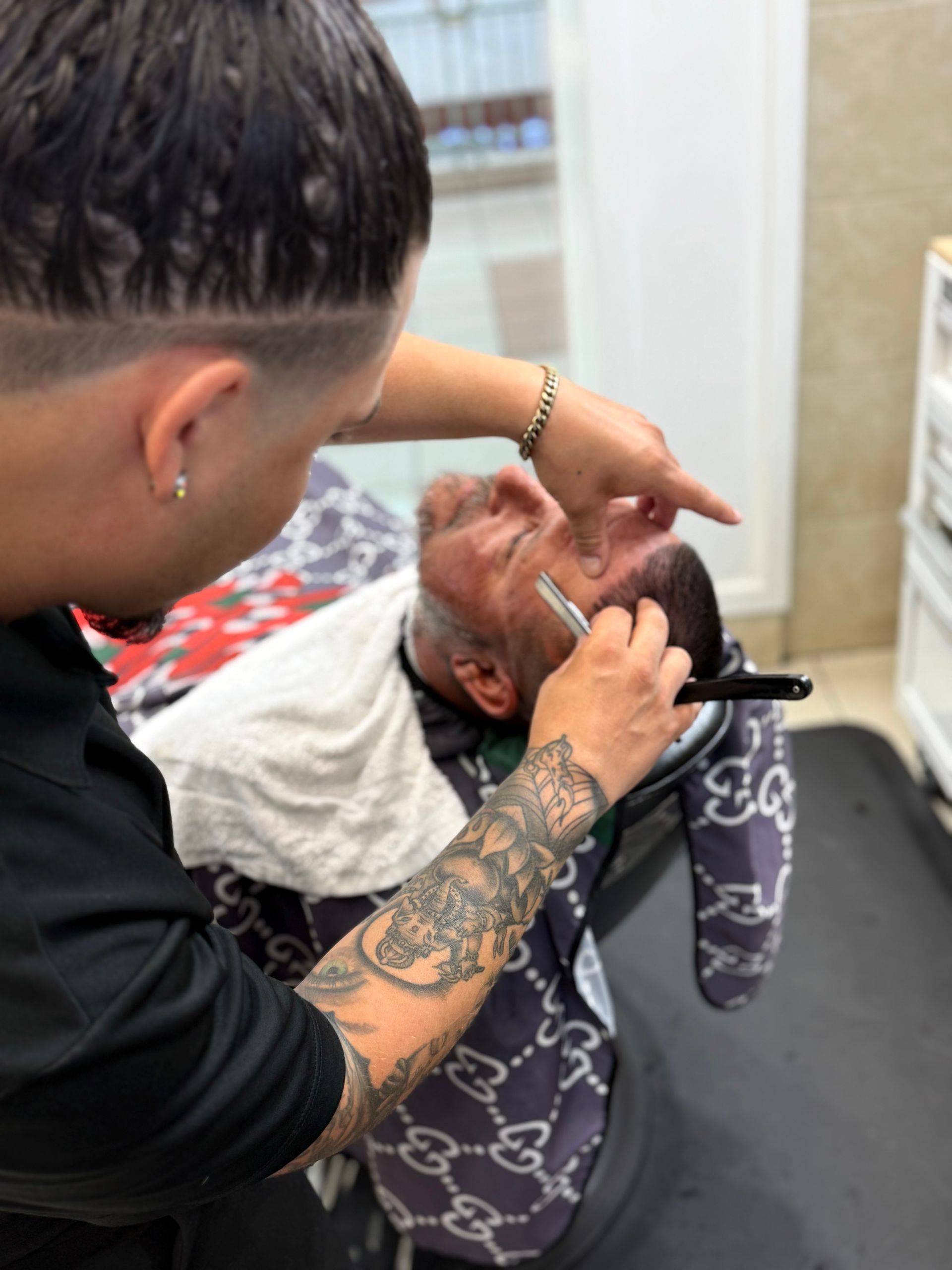 Barber shaving a man's face with a straight razor. The man is lying back, eyes closed. The barber has tattoos and a detailed haircut.