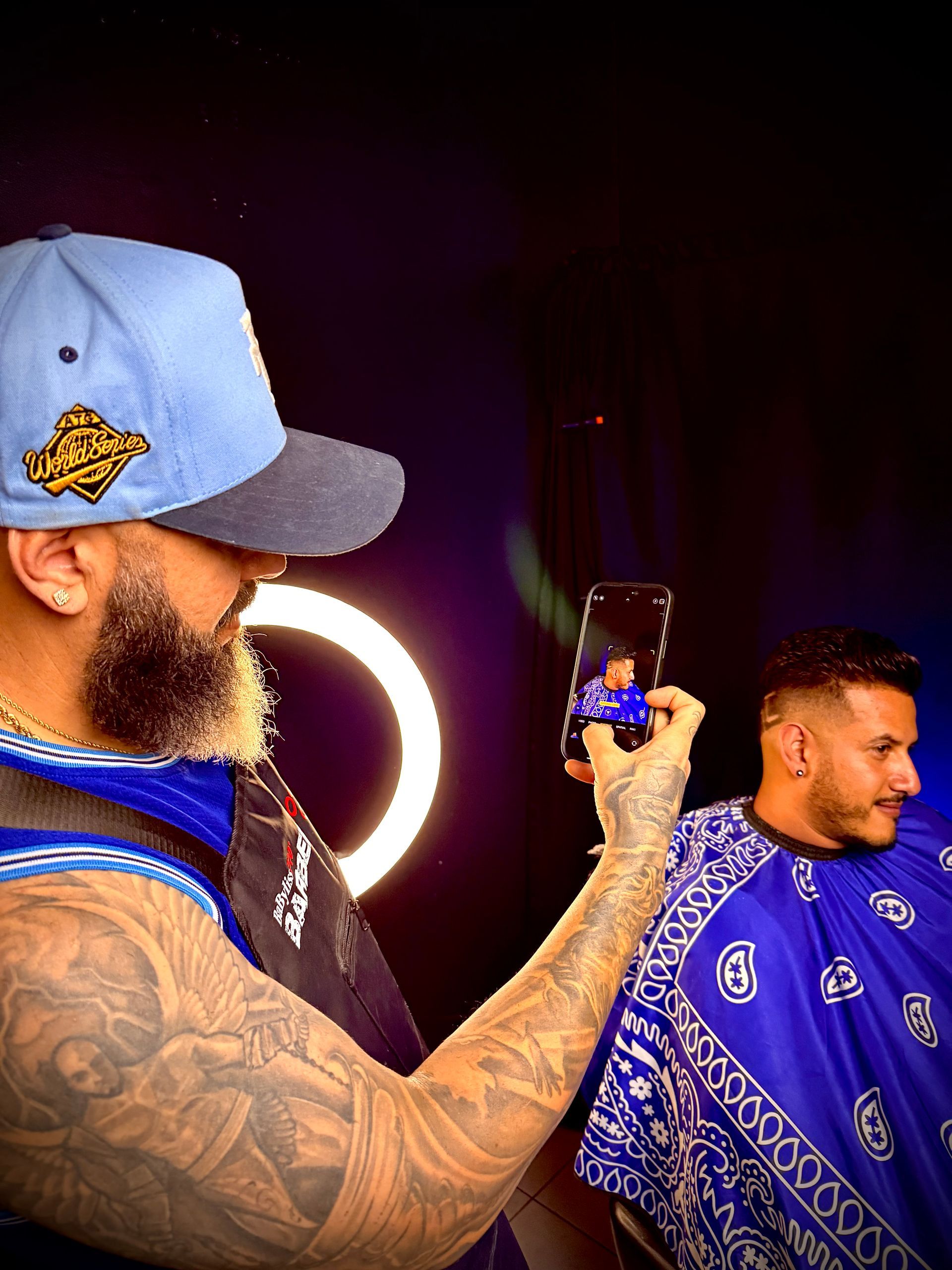 Barber taking a photo of a client's haircut; both men in blue, bright ring light in background.