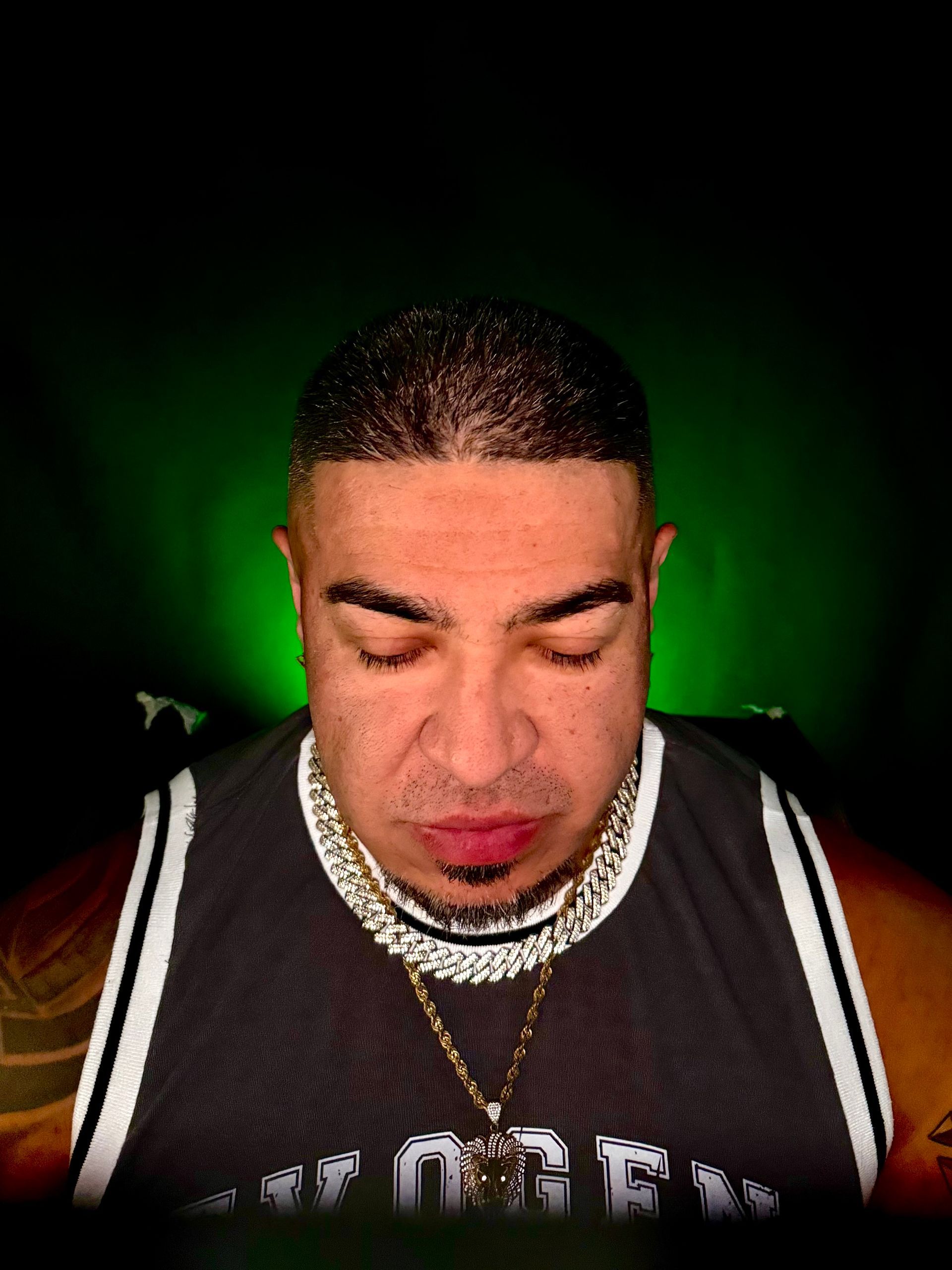 Man wearing a tank top and multiple gold chains, looking down with a contemplative expression. Green background.