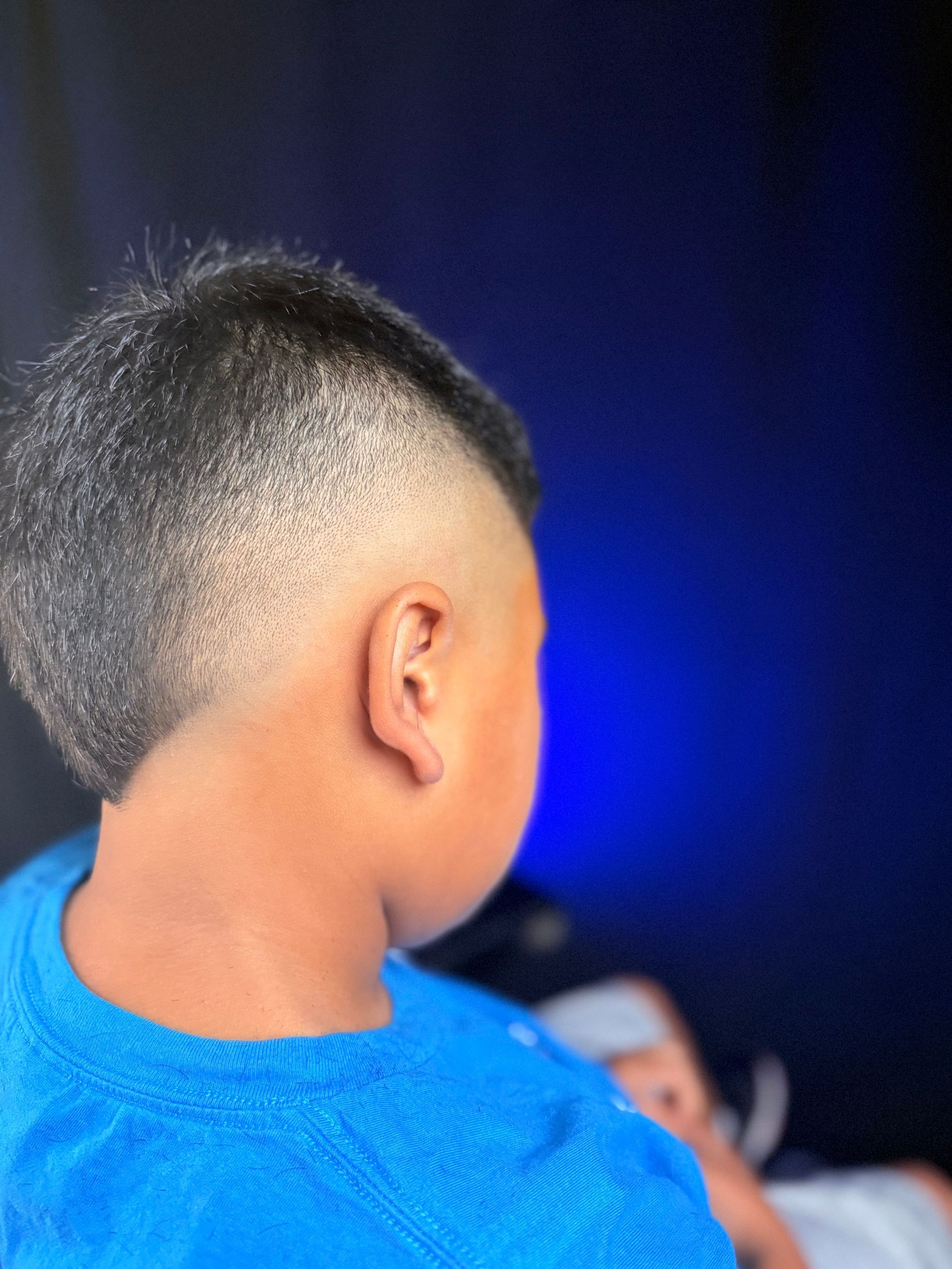 Boy with a faded haircut, wearing a blue shirt, facing away, in front of a dark blue background.
