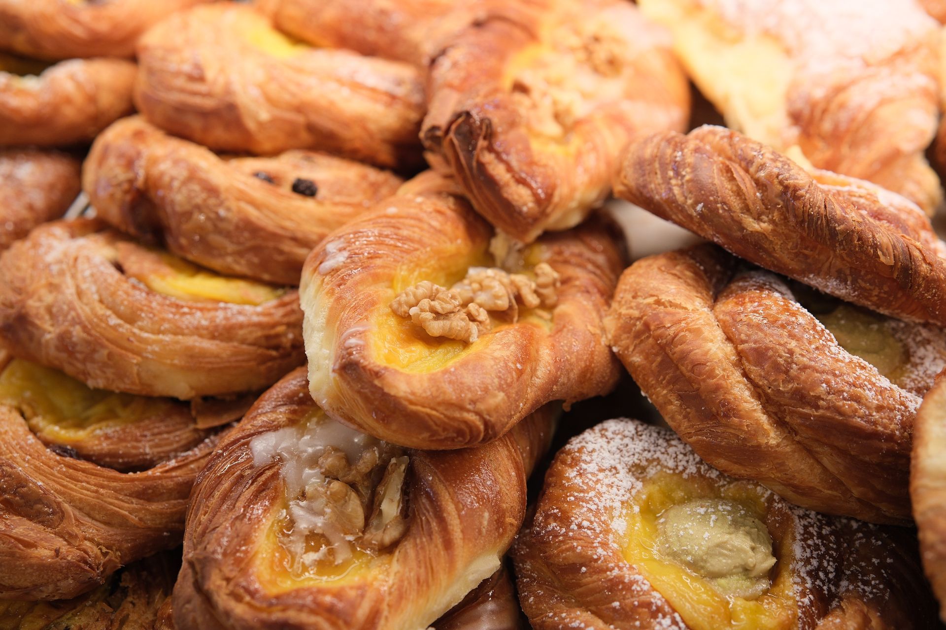 Pasticcini assortiti: danesi dorati, alcuni con ripieno di crema pasticcera, noci e zucchero a velo, ammucchiati insieme.