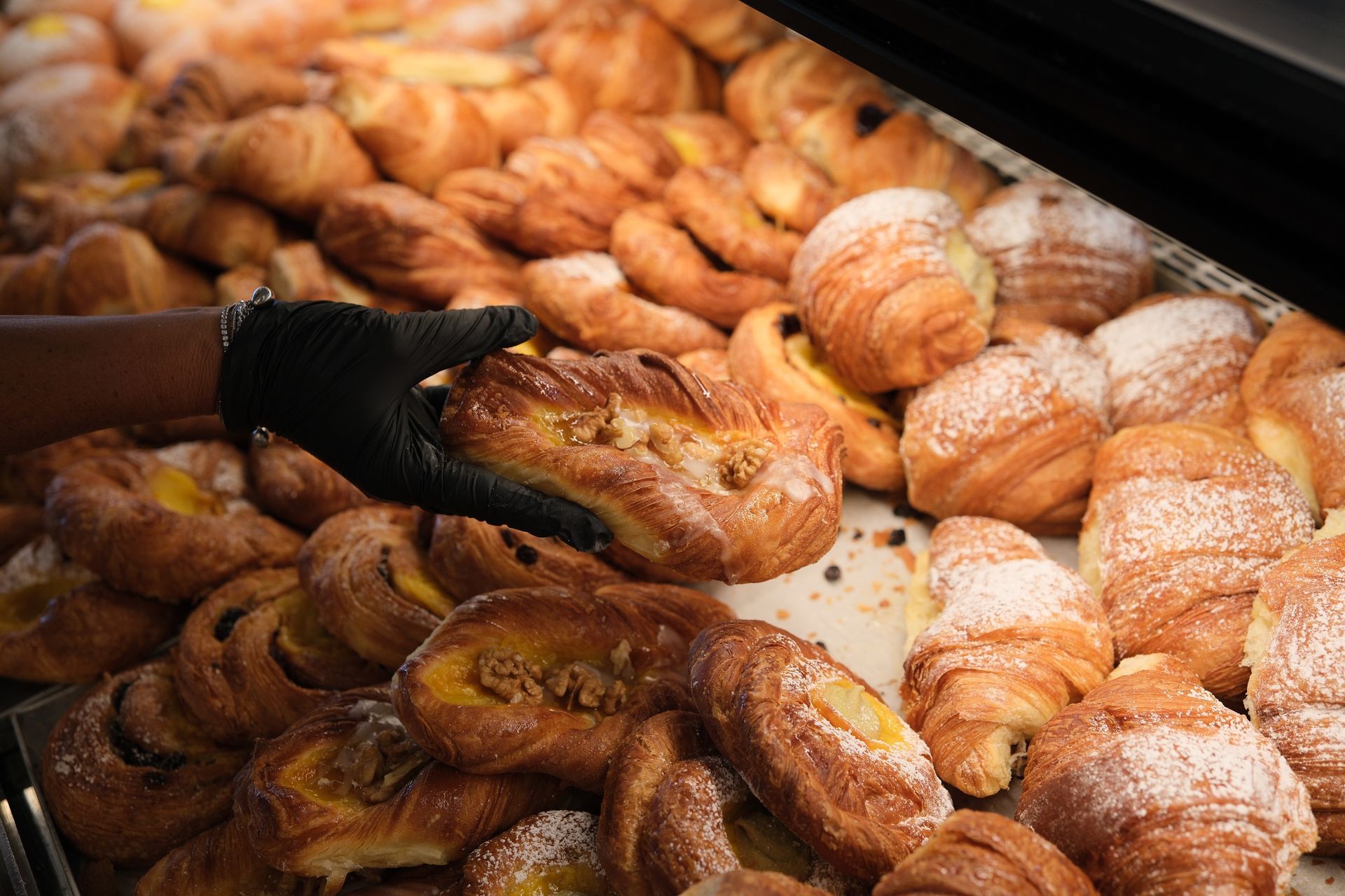 Mano che regge un dolce in una vetrina da forno piena di vari prodotti da forno, come i croissant.