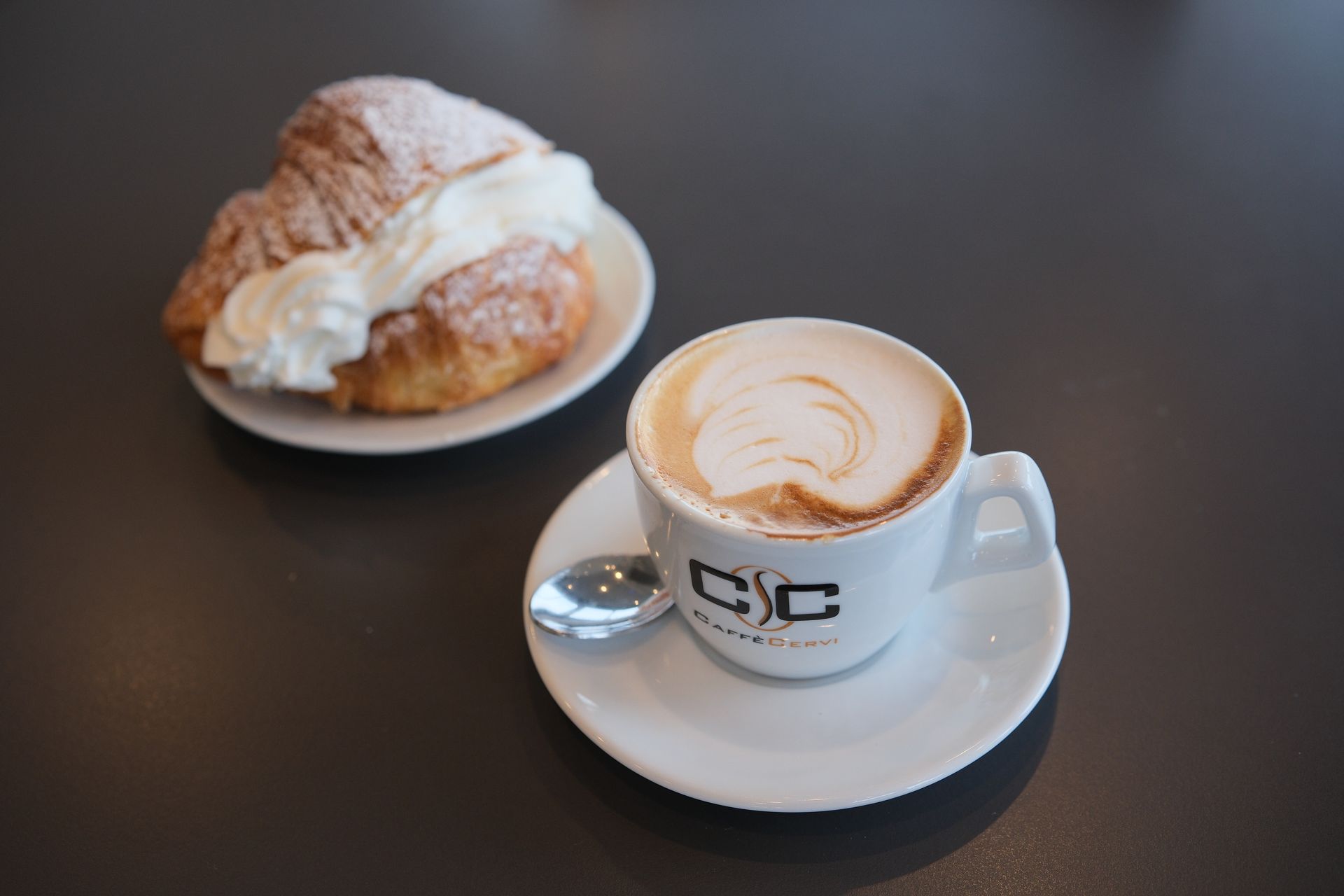 Tazza di cappuccino e croissant con panna montata su un piattino su un tavolo buio.