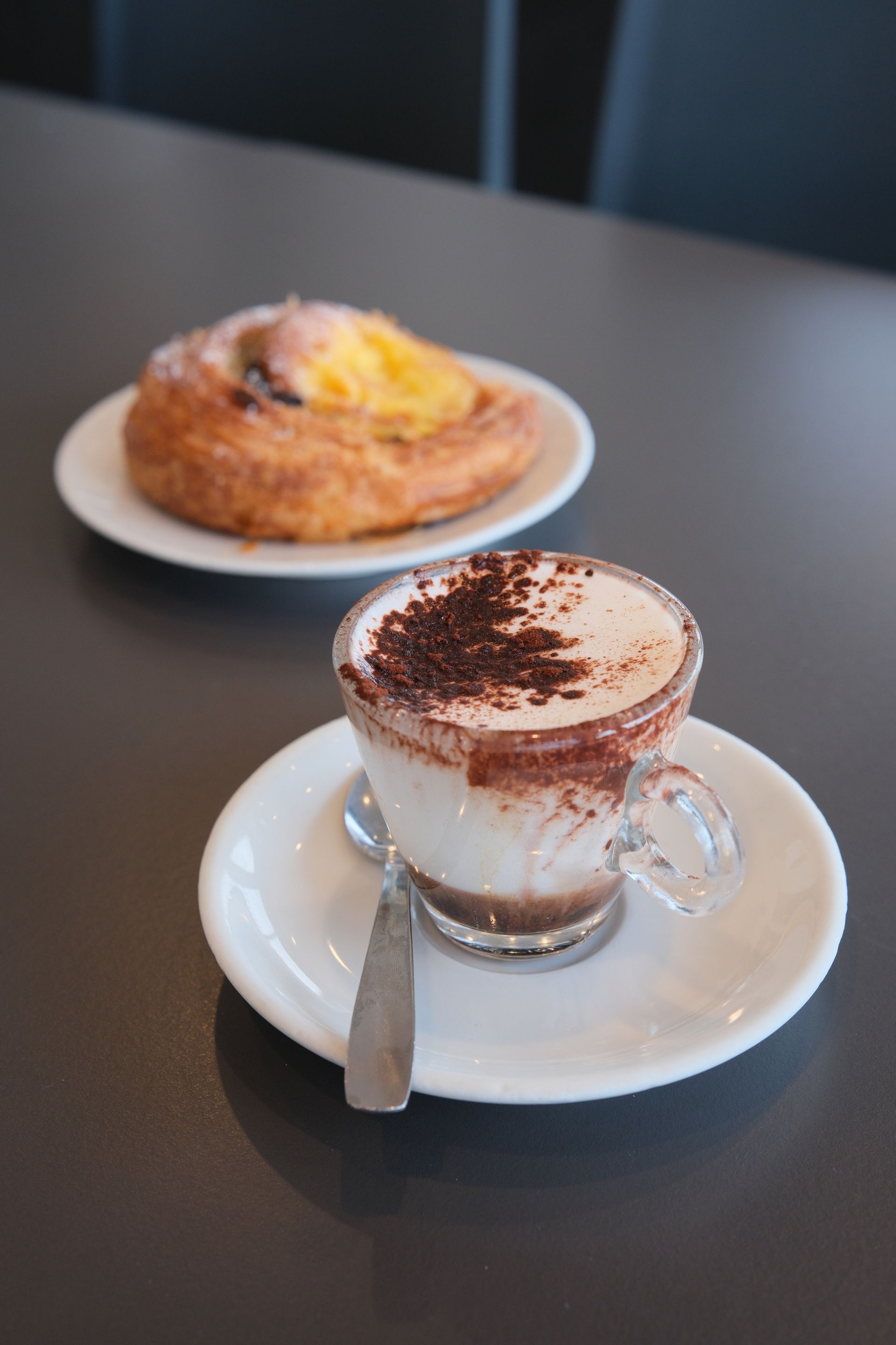 Tazza di cioccolata calda ricoperta di polvere di cioccolato accanto a un dolce su un piatto bianco.