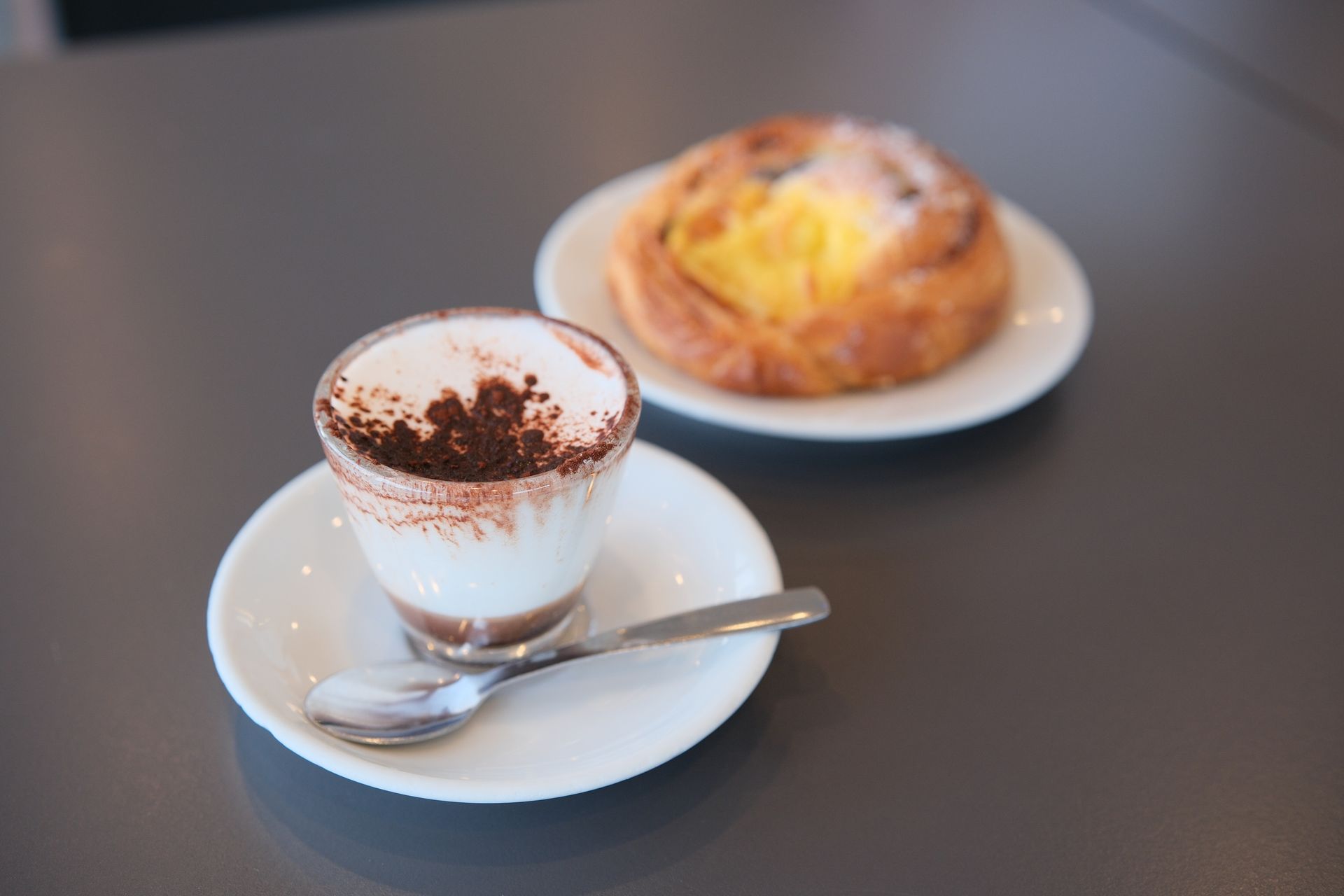 Bevanda al caffè con cacao e pasticcini su un piattino e un piatto bianchi, su un tavolo grigio.