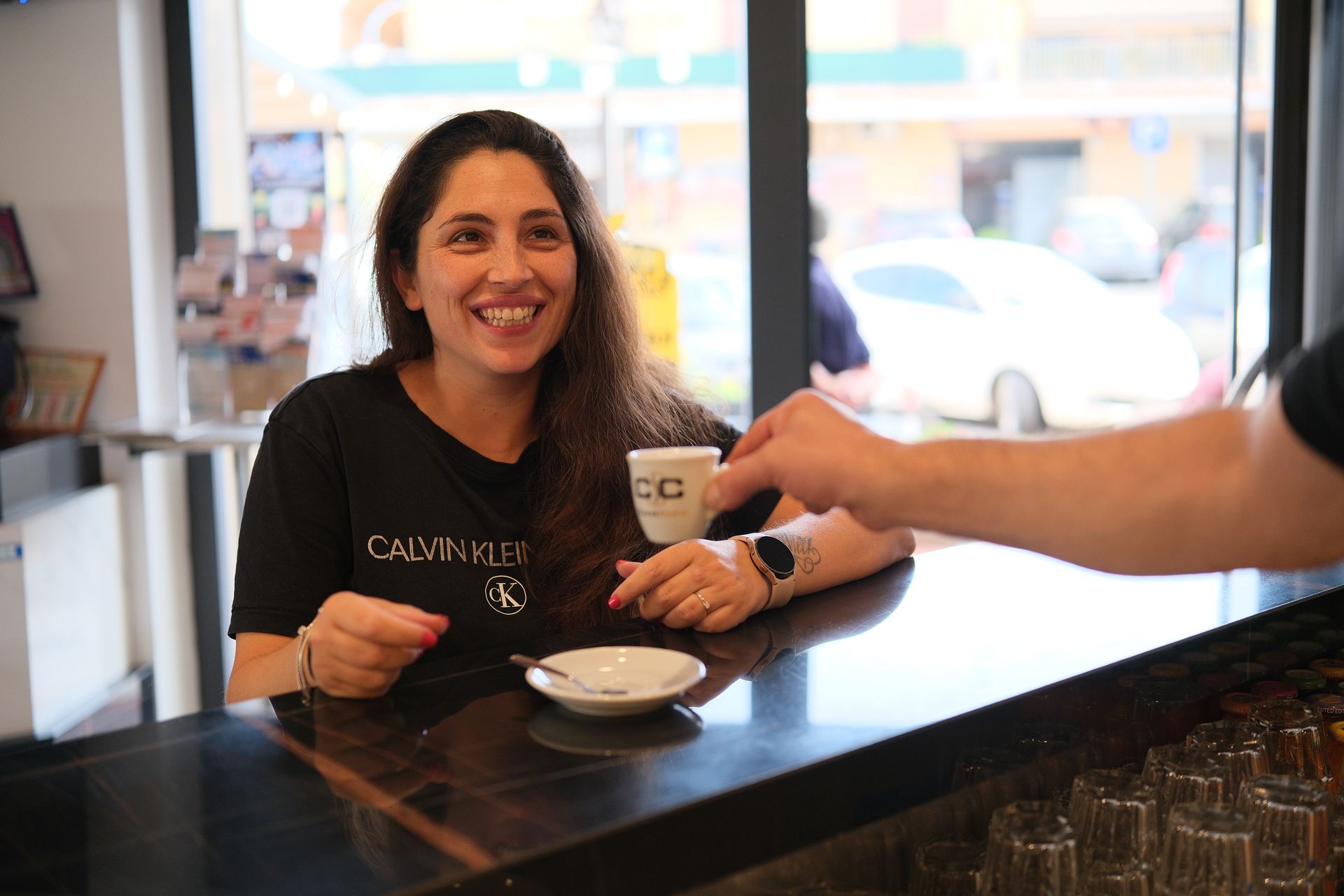 Una donna al bancone di un bar sorride mentre una mano le offre una piccola tazza di caffè.