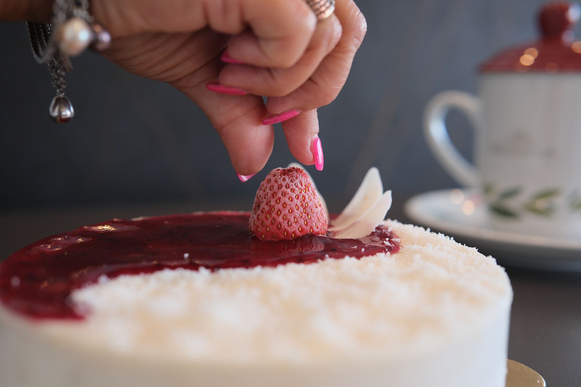 Mano che mette una fragola su una torta bianca con glassa rossa. Tazza sullo sfondo.