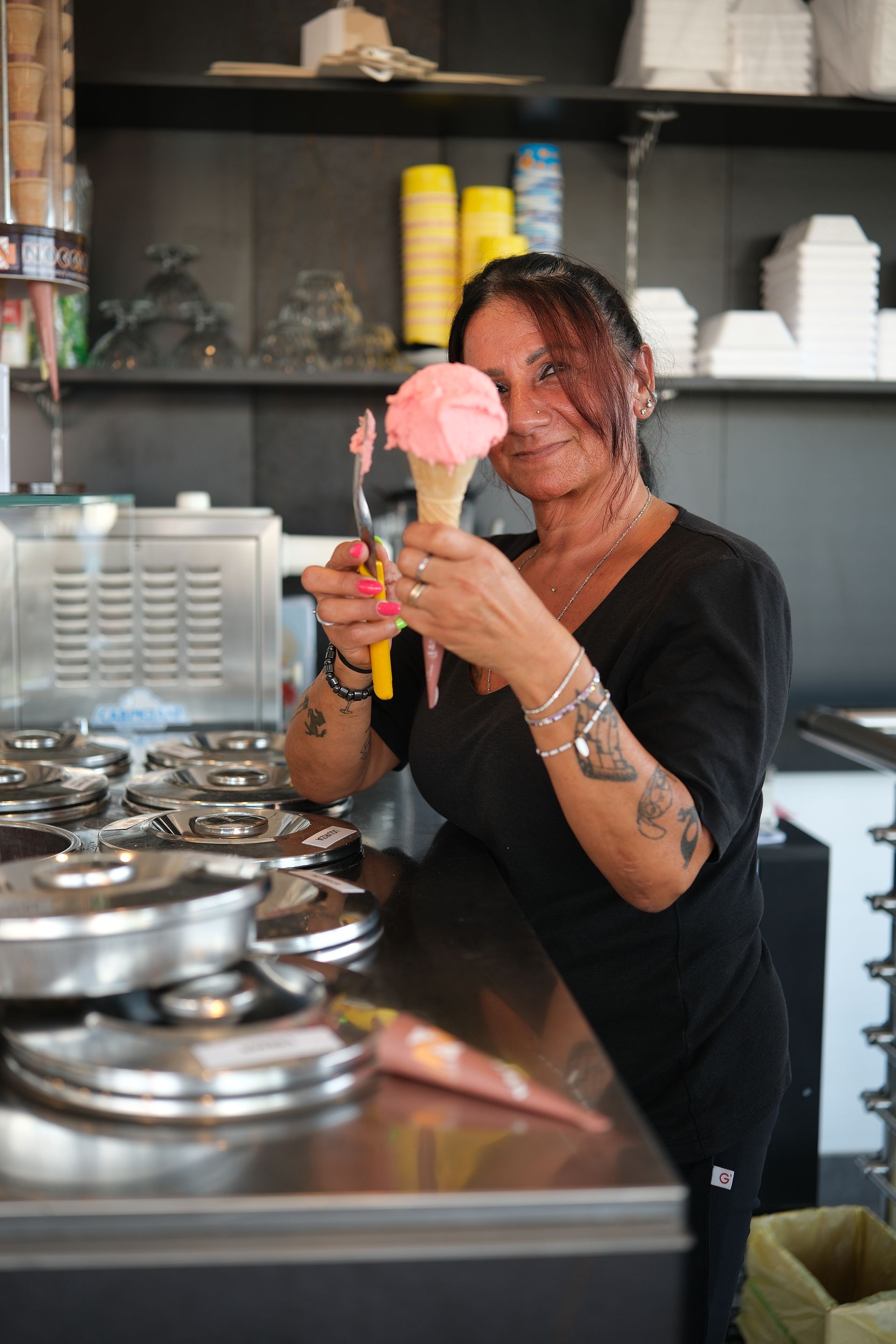 Una donna con i tatuaggi tiene in mano un cono gelato rosa e sorride in una gelateria.