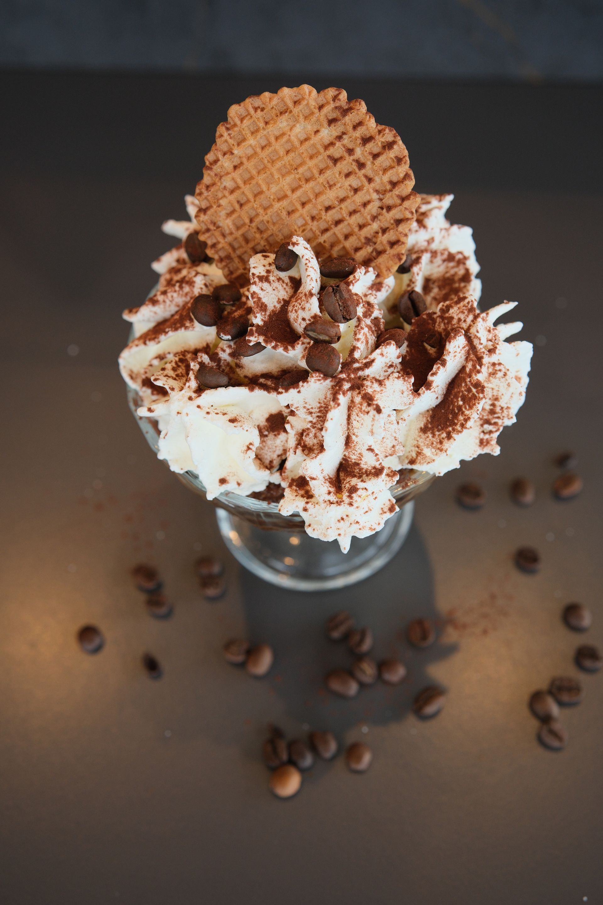 Dessert con panna montata, biscotti, scaglie di cioccolato e chicchi di caffè su una superficie scura.