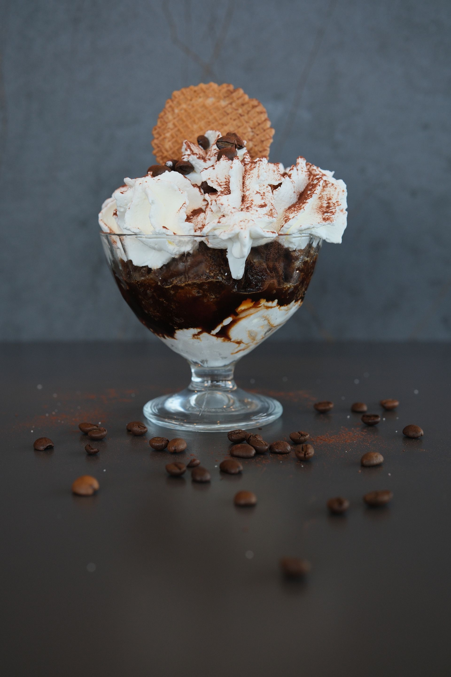 Dessert al caffè servito in un bicchiere, guarnito con panna montata, cacao in polvere, una cialda e chicchi di caffè.