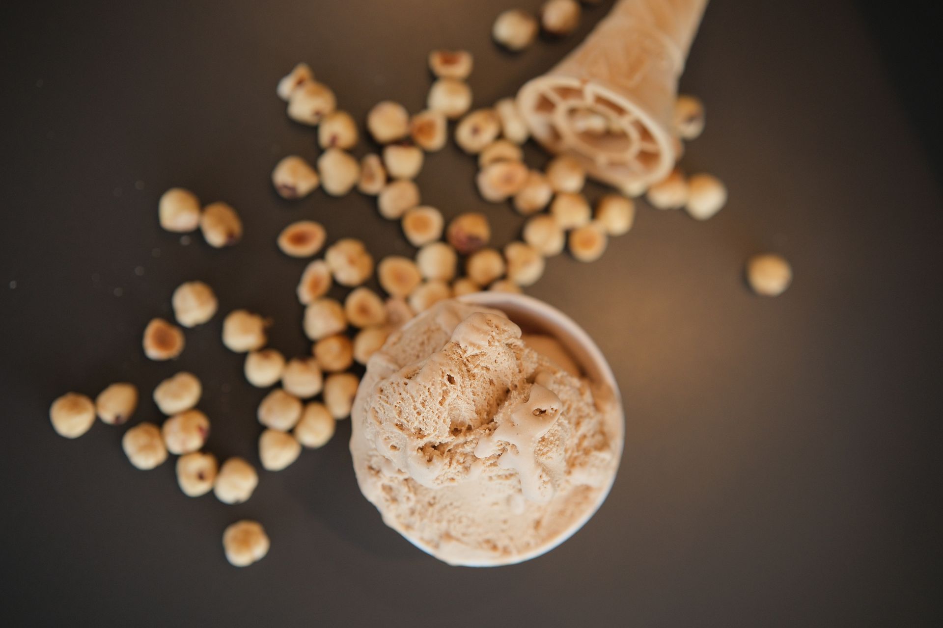 Gelato in una coppetta bianca, con nocciole e un cono su una superficie scura.