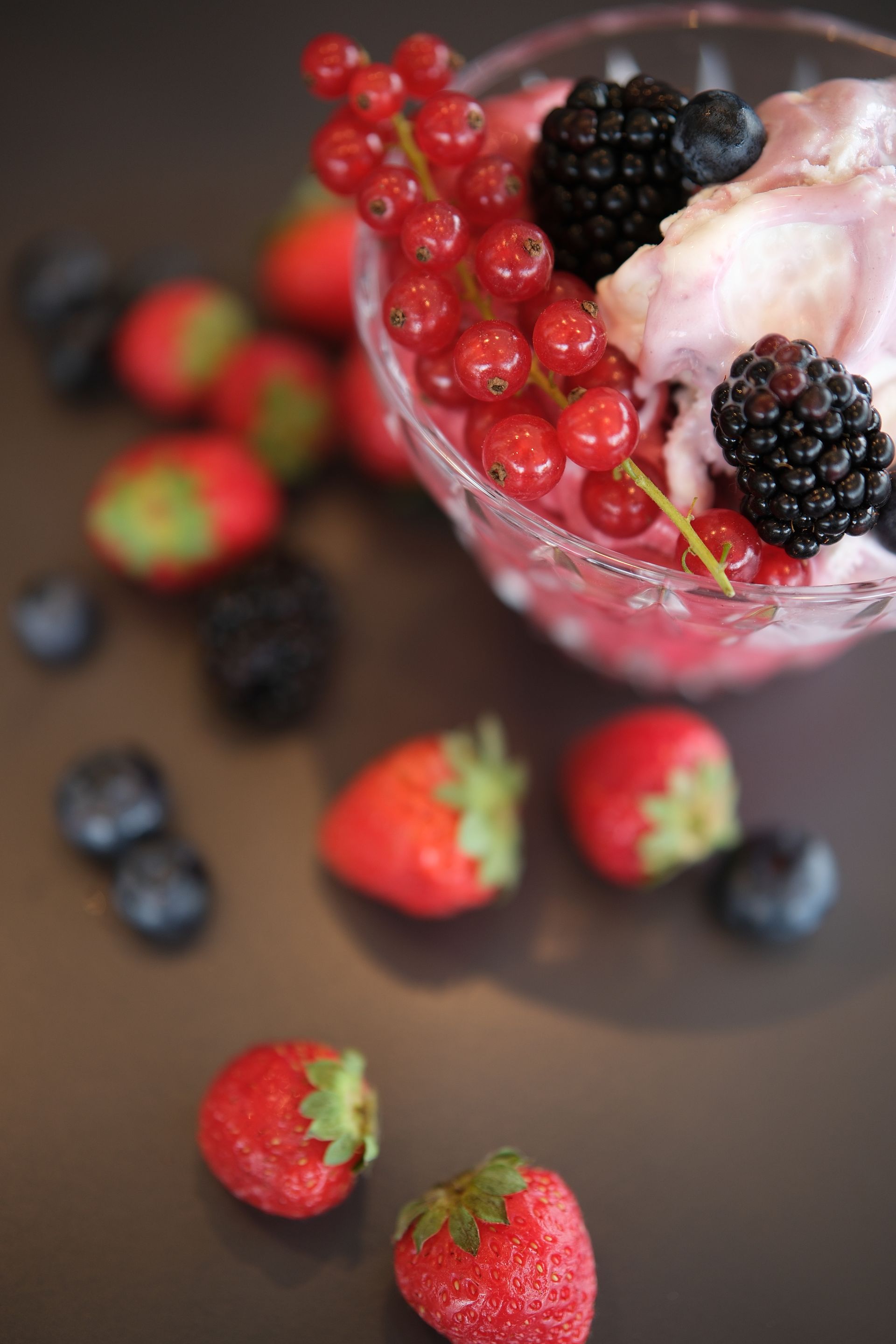 Ciotola di vetro con dessert rosa, ribes rossi e more sparsi intorno.