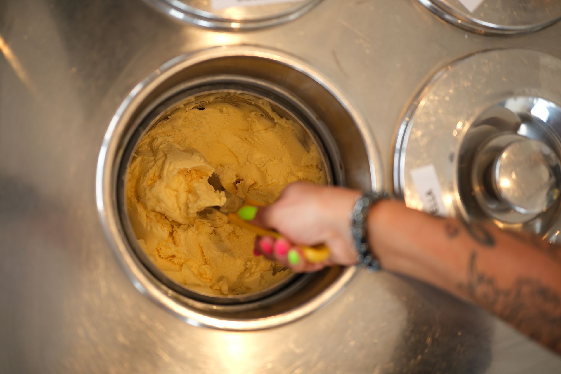Persona che preleva un gelato giallo da un contenitore di metallo con un cucchiaio dal manico colorato.