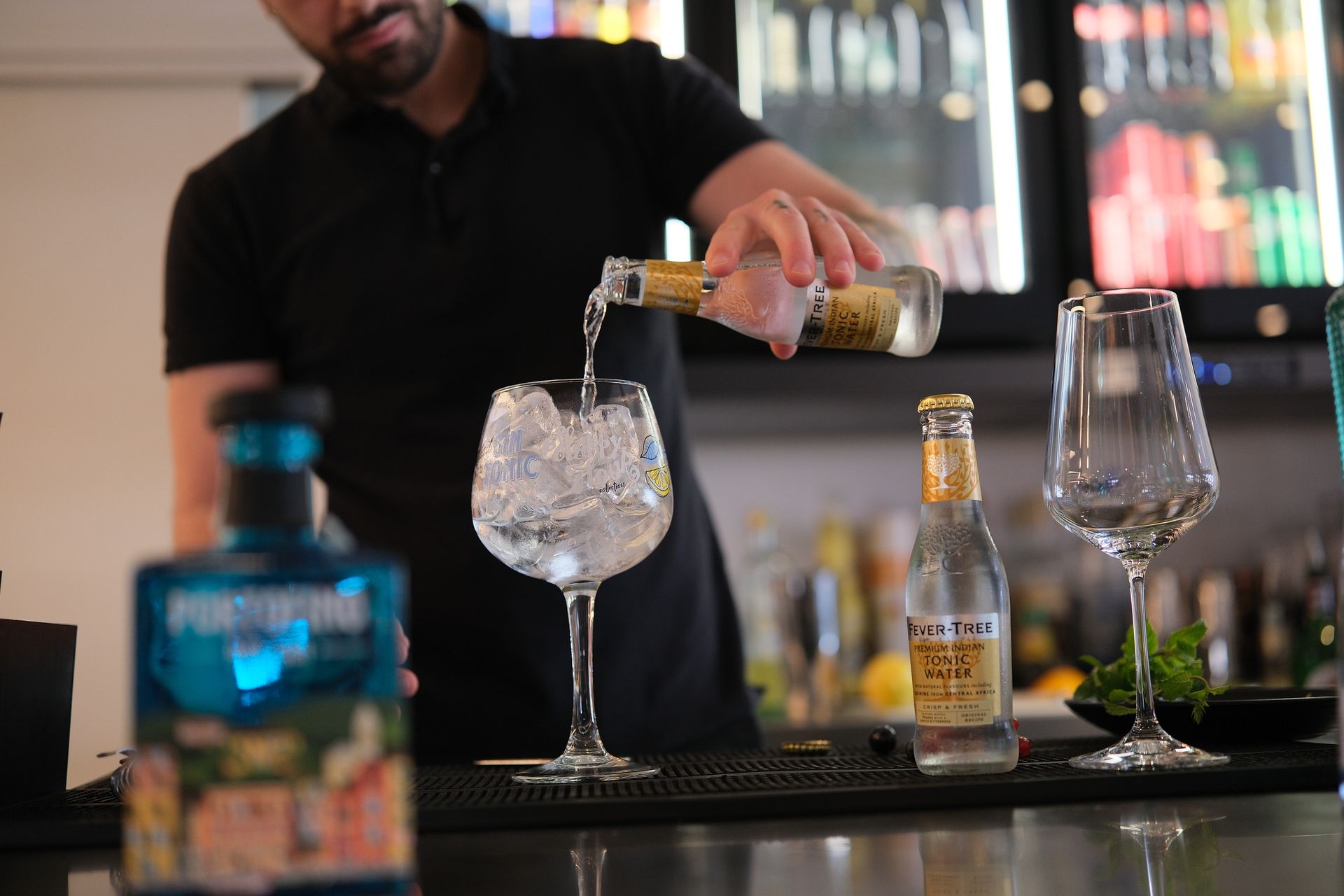 Barista che versa un drink in un bicchiere con ghiaccio in un bar; bottiglie di acqua tonica e gin.