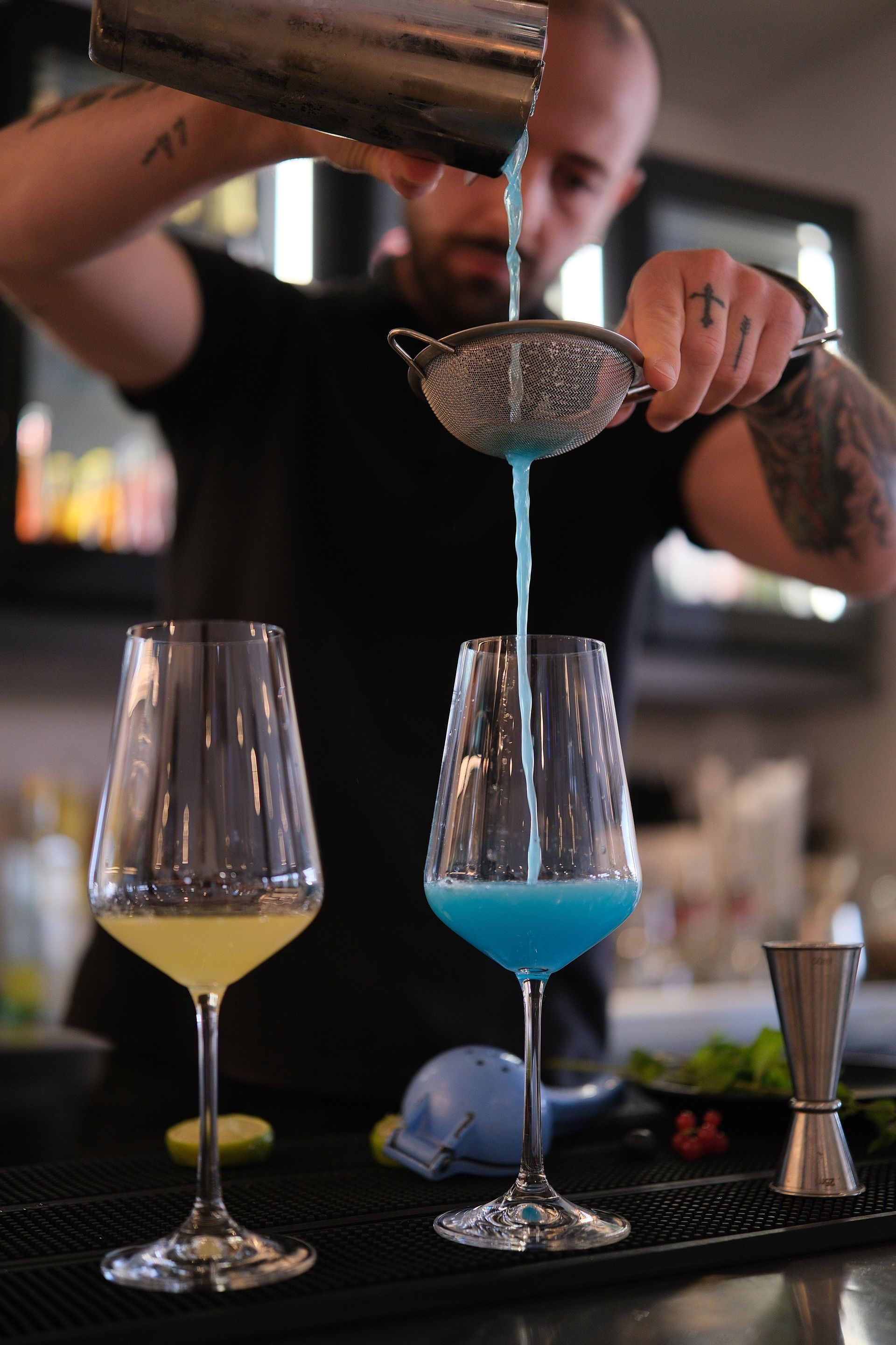Barista che versa un cocktail blu in un bicchiere, un altro bicchiere con un drink giallo, ambiente da bar.
