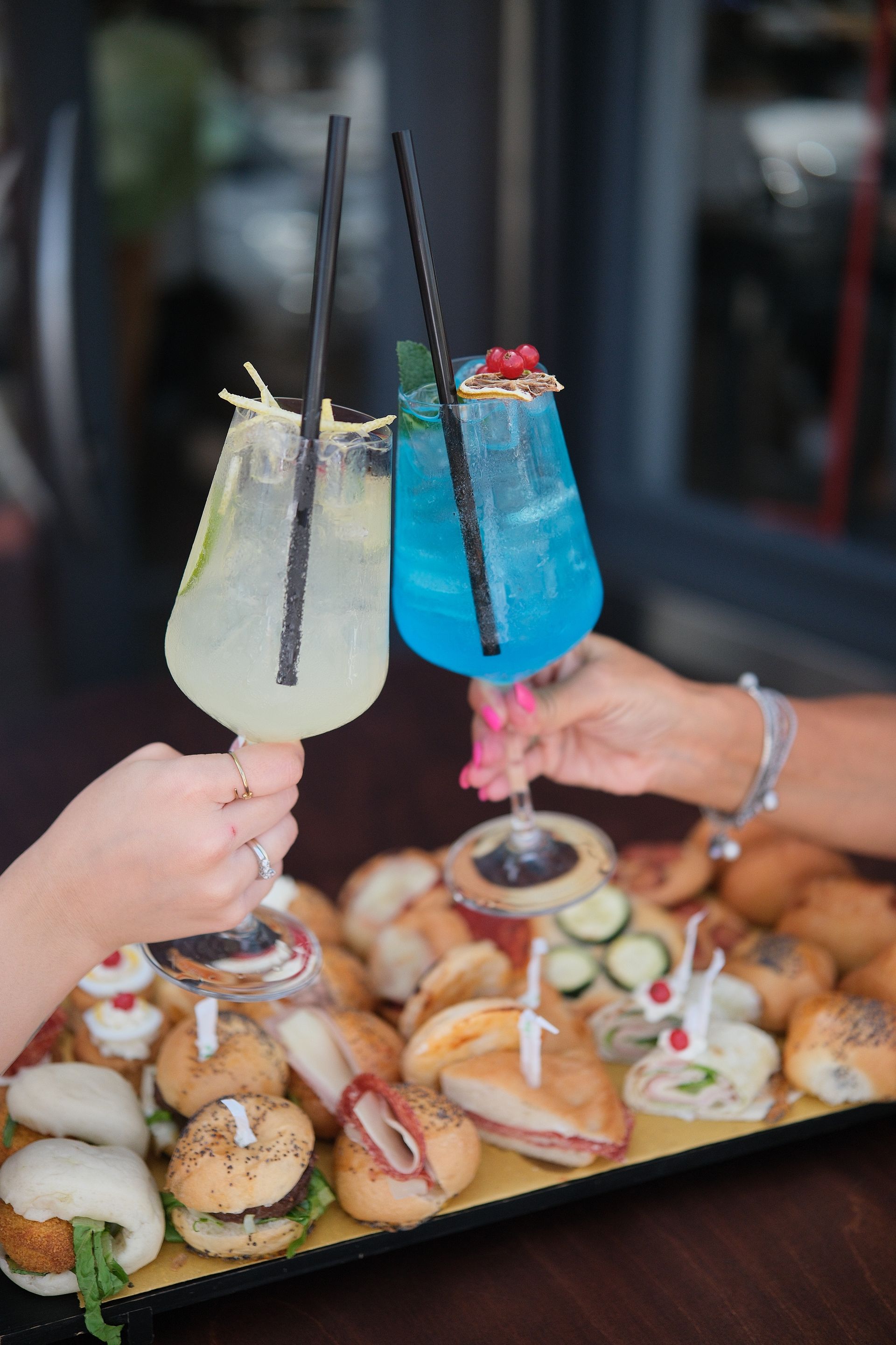 Due mani che brindano a un cocktail con un vassoio di cibo: drink giallo chiaro e blu, ambientazione all'aperto.