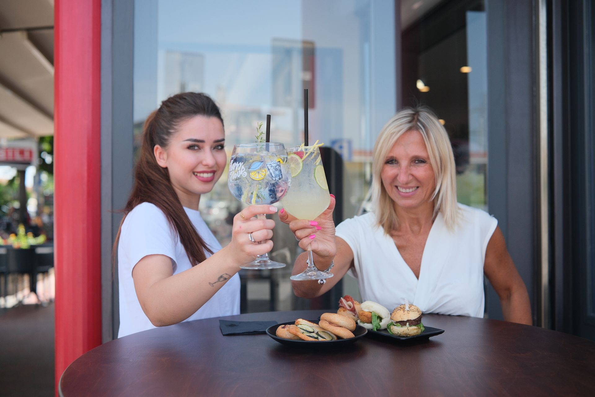 Due donne sorridono e brindano all'aperto. Tavolo con stuzzichini, sullo sfondo un ristorante.