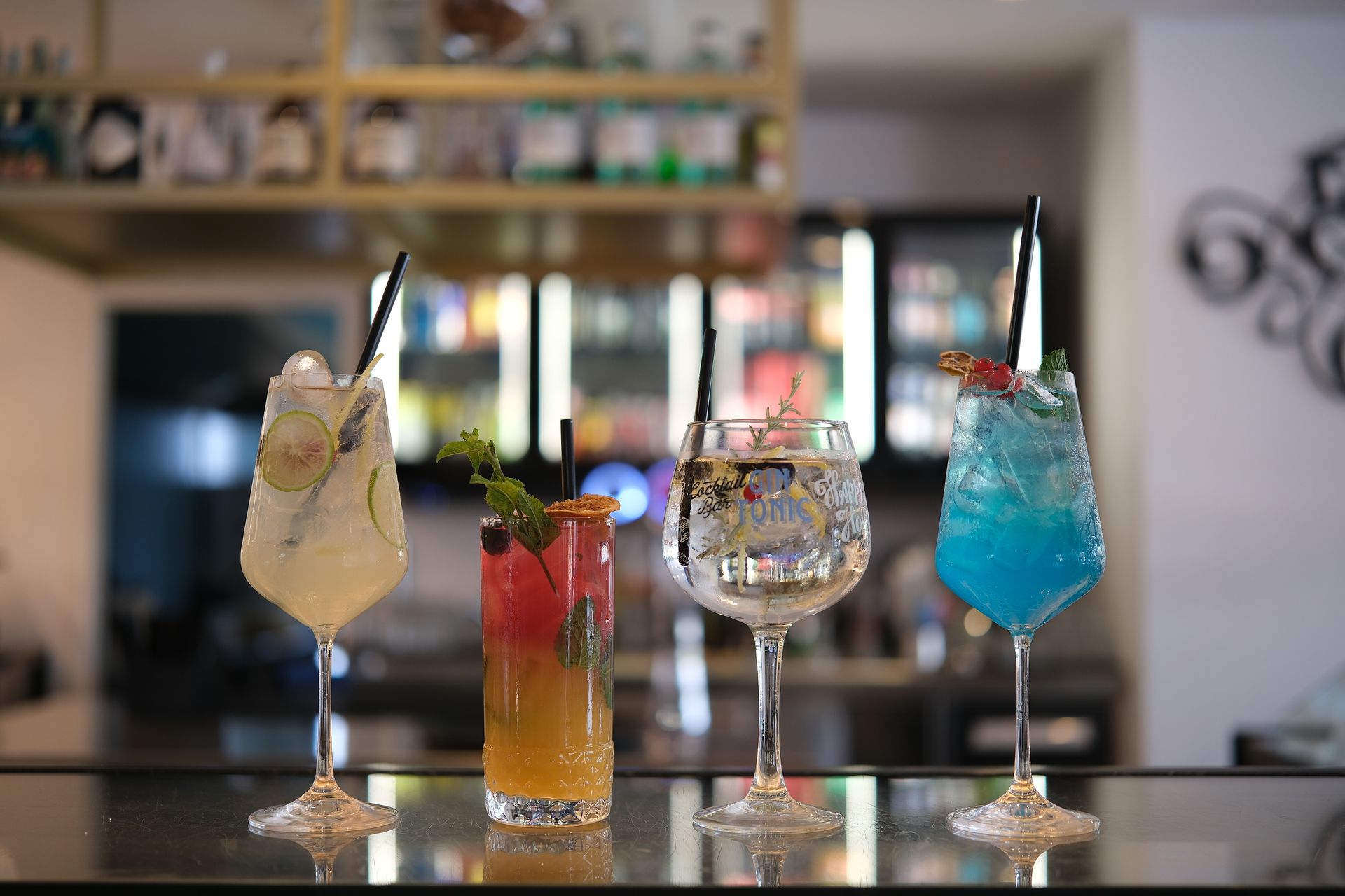 Quattro cocktail colorati sul bancone di un bar, ognuno con una cannuccia, sullo sfondo sfocato del bar.