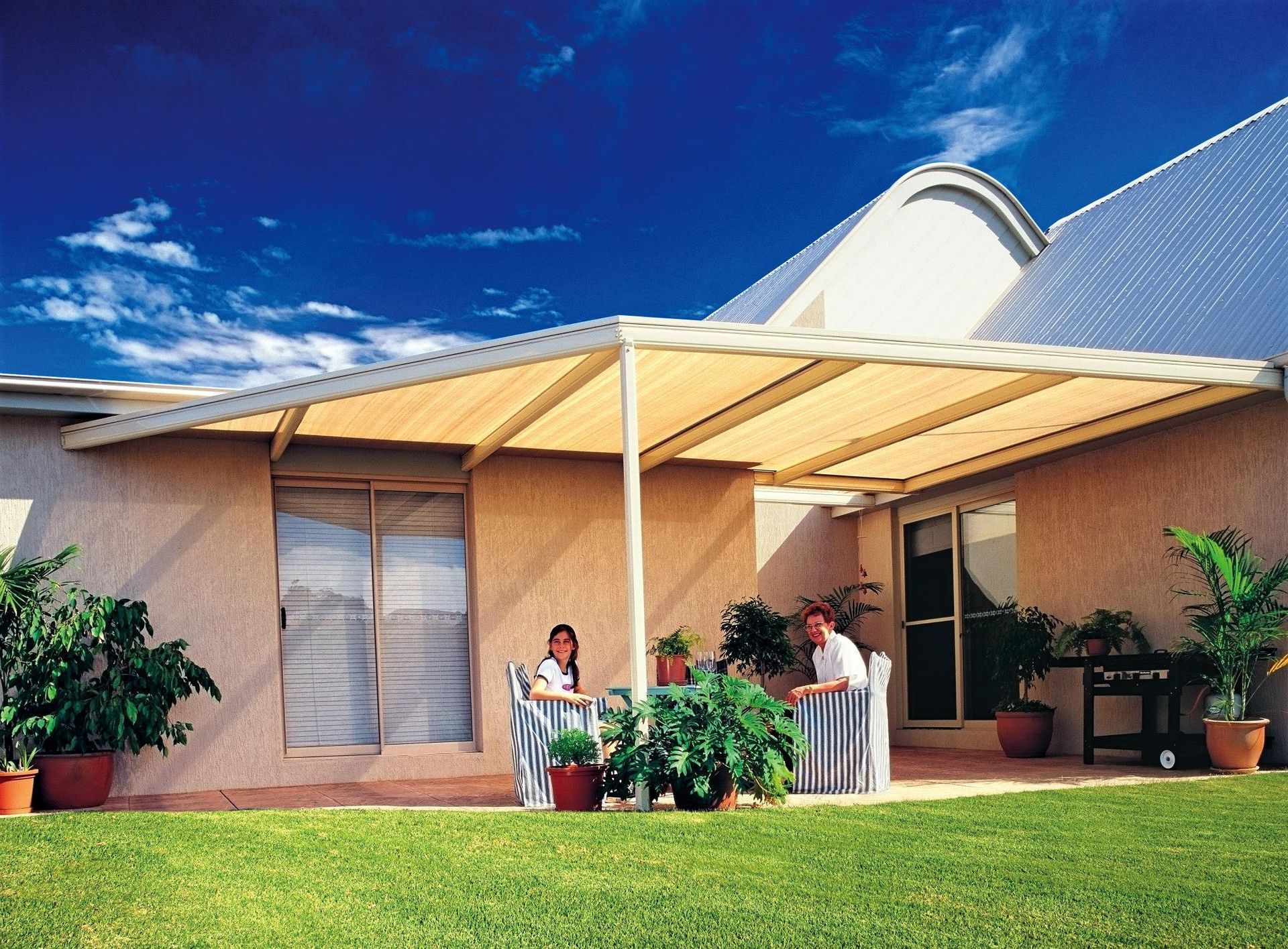 Two people sit under a canopy on a patio in front of a house
