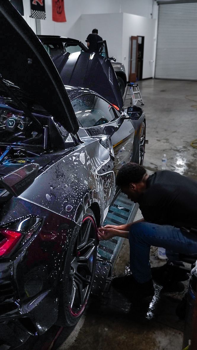 Man working on a wet, dark blue sports car in a garage. Car's hood open.