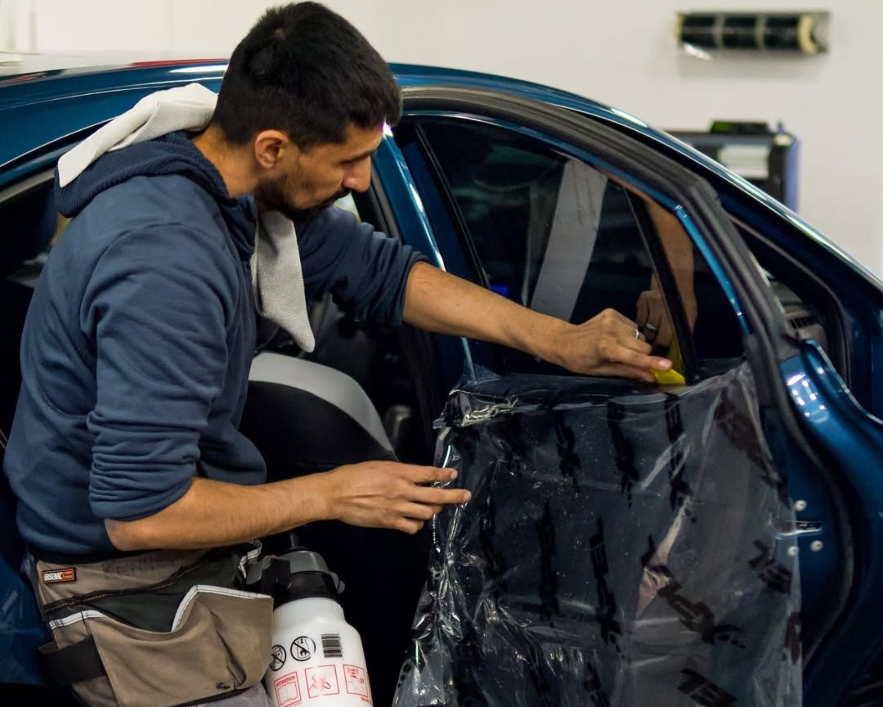 Person applying tint to a car window in a garage. They are holding tint film, with a spray bottle attached to a waist belt.