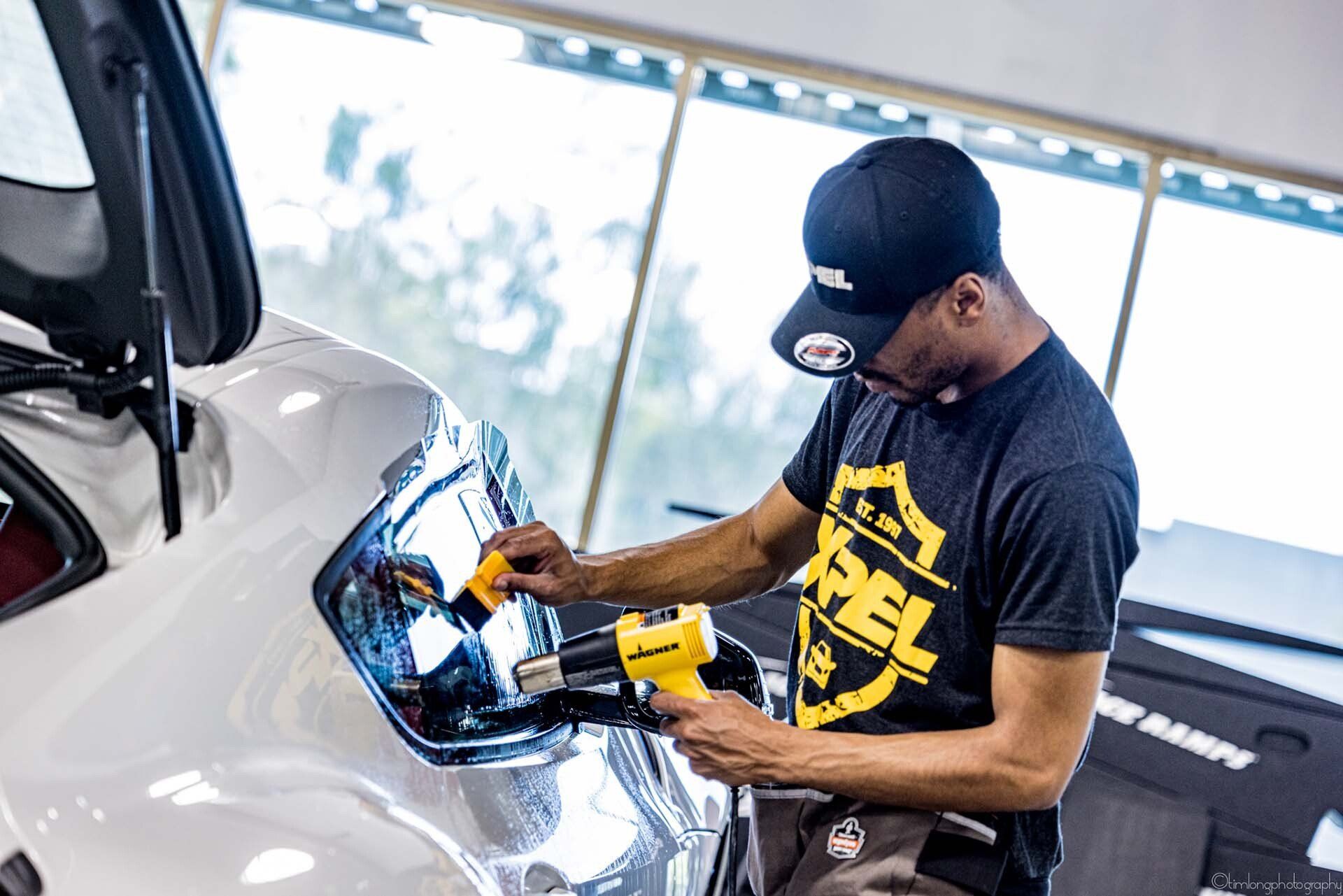 Man applying tint to a car window with a heat gun. Wearing a baseball cap and working inside.