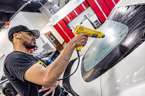 Man using a heat gun on a car window to apply tint in a garage.