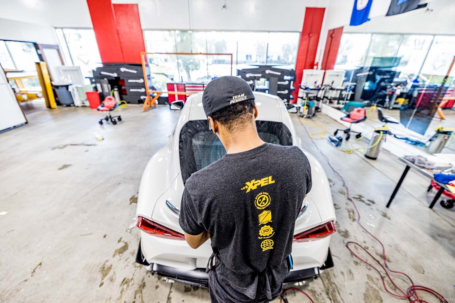 Man applying tint to a white car's rear window in a workshop. He's wearing a hat and black shirt.