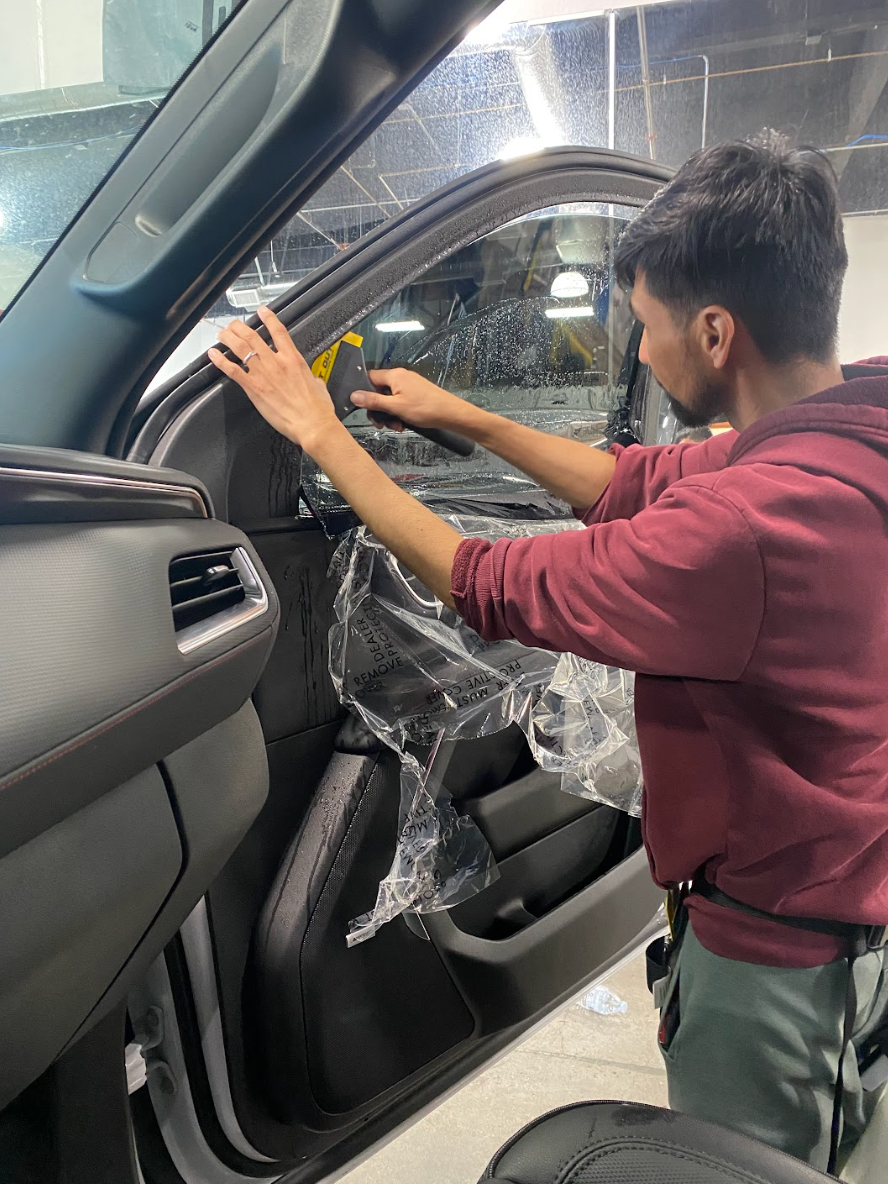 Person applying tint to car window in a garage. Wearing red hoodie. Window is partially covered in plastic.