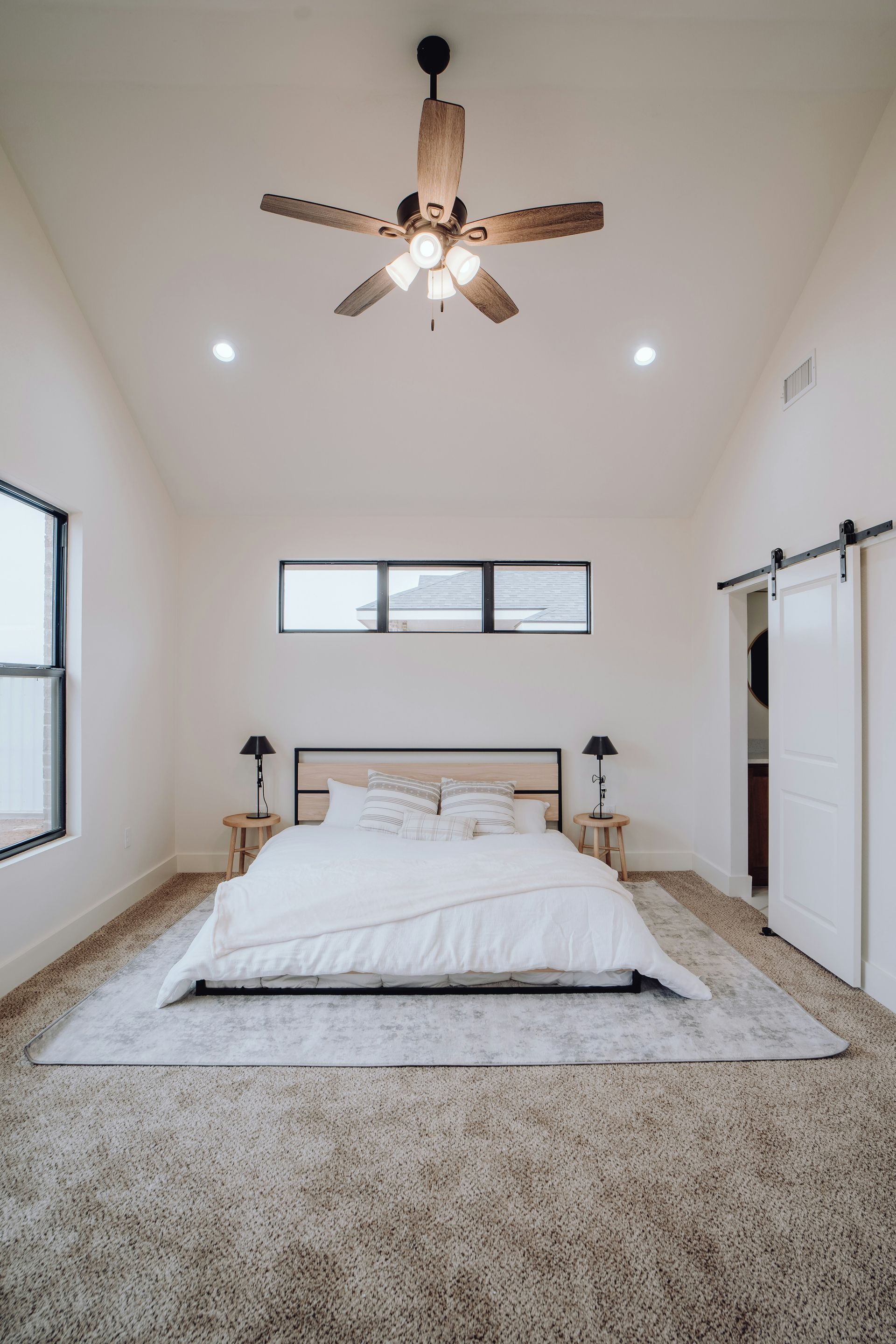 Bedroom with vaulted ceiling, light walls, bed, rug, windows, barn door, and ceiling fan.