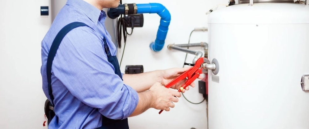 A plumber in a blue shirt and suspenders uses red pliers on a water heater.