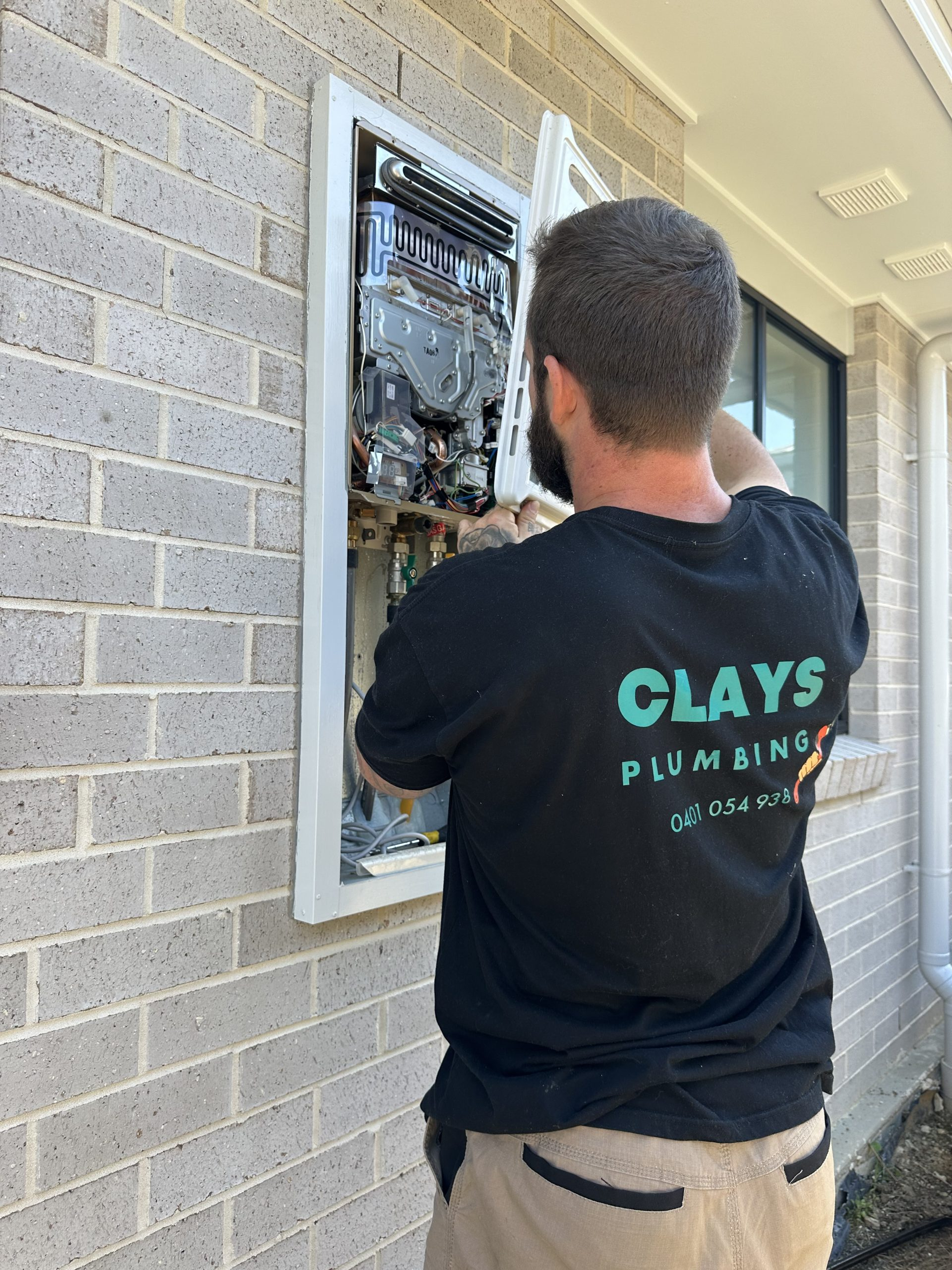 Plumber in black t-shirt installs a gas water heater on a brick wall.