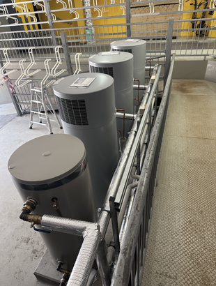 Four large, gray water heaters in a utility room, adjacent to a metal structure and wall.