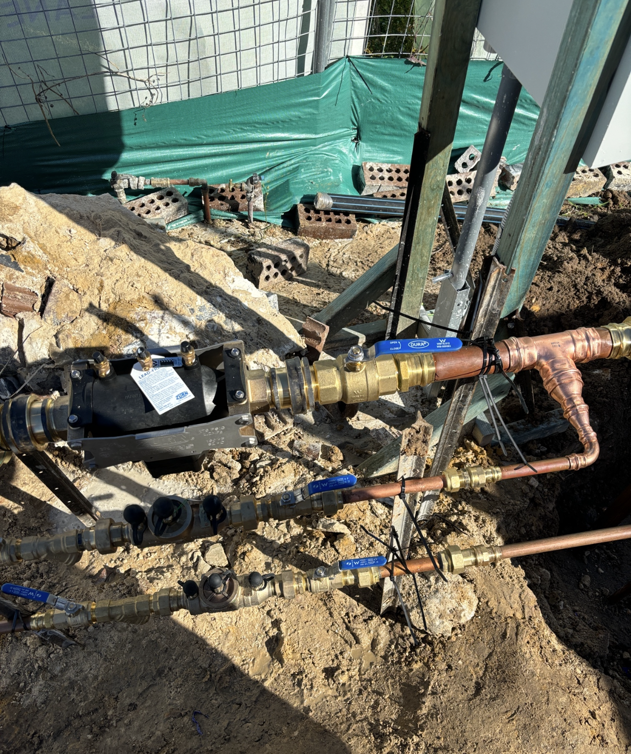 Plumbing pipes and valves laid in a dirt trench, near a metal support beam and brick.